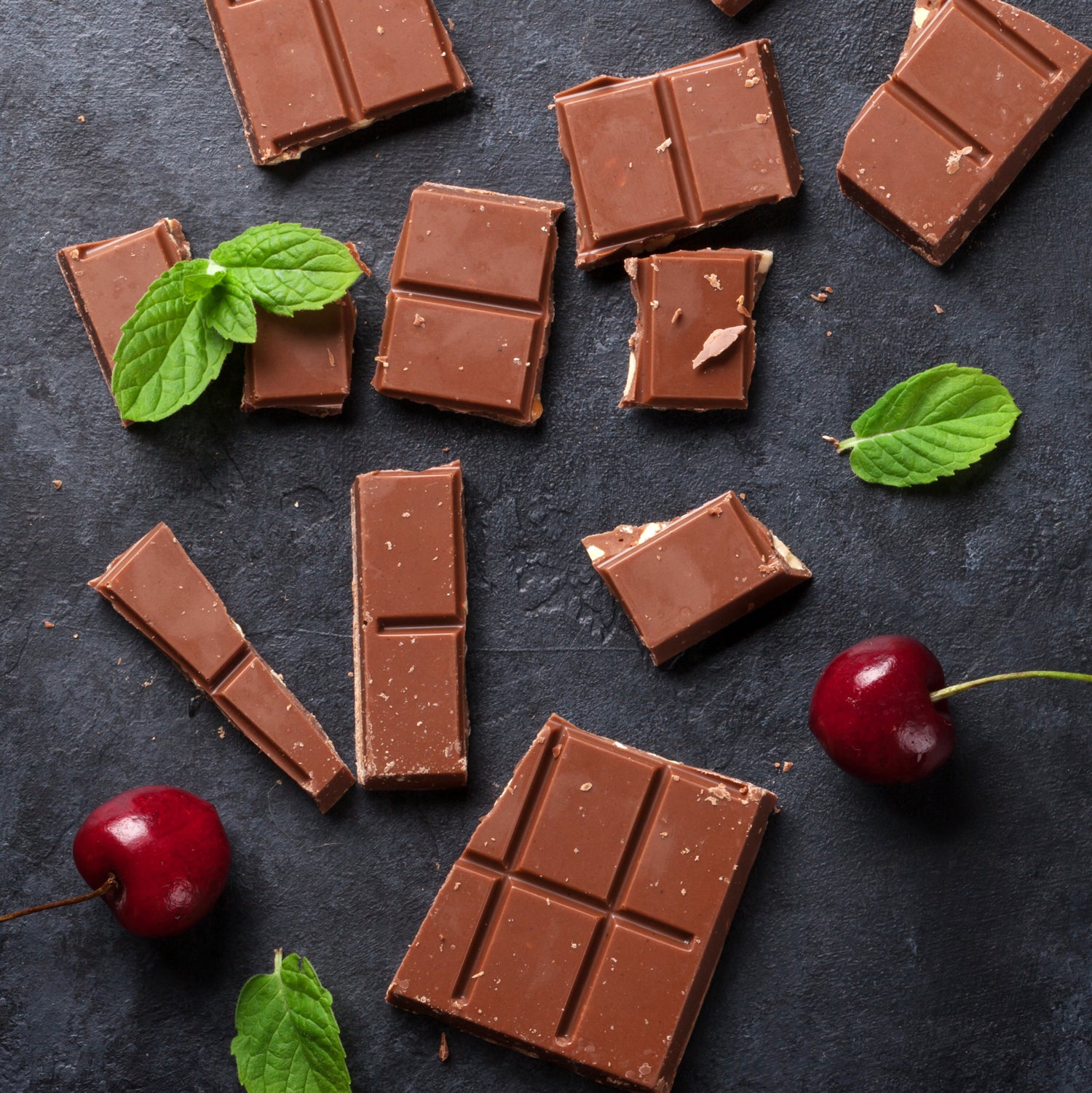 Assorted milk chocolate pieces with fresh mint leaves and Tuscany Candle® EVD Sweet Cherry Long-Lasting Scented Jar Candle (14 oz) on a dark textured background.