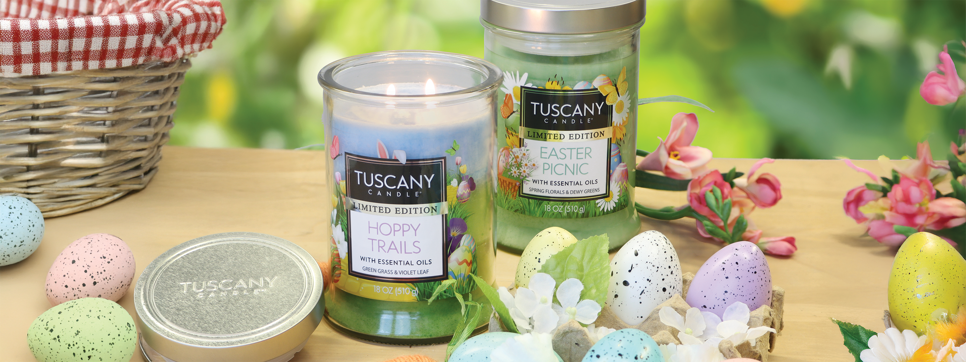 Two Tuscany candles labeled "Hoppy Trails" and "Easter Picnic" are on a table with decorated eggs and flowers. A wicker basket is in the background.