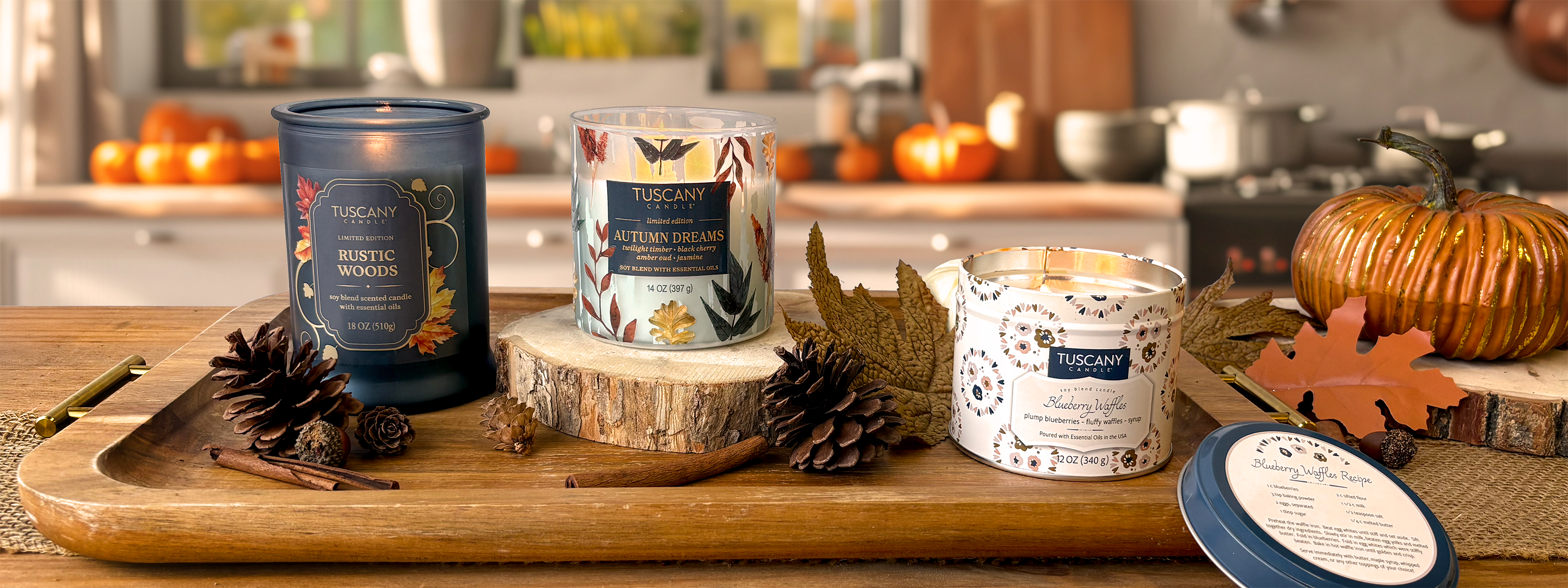 Three autumn-themed Tuscany Candle jars, pinecones, cinnamon sticks, and a pumpkin are arranged on a wooden tray in a kitchen with fall decorations in the background.