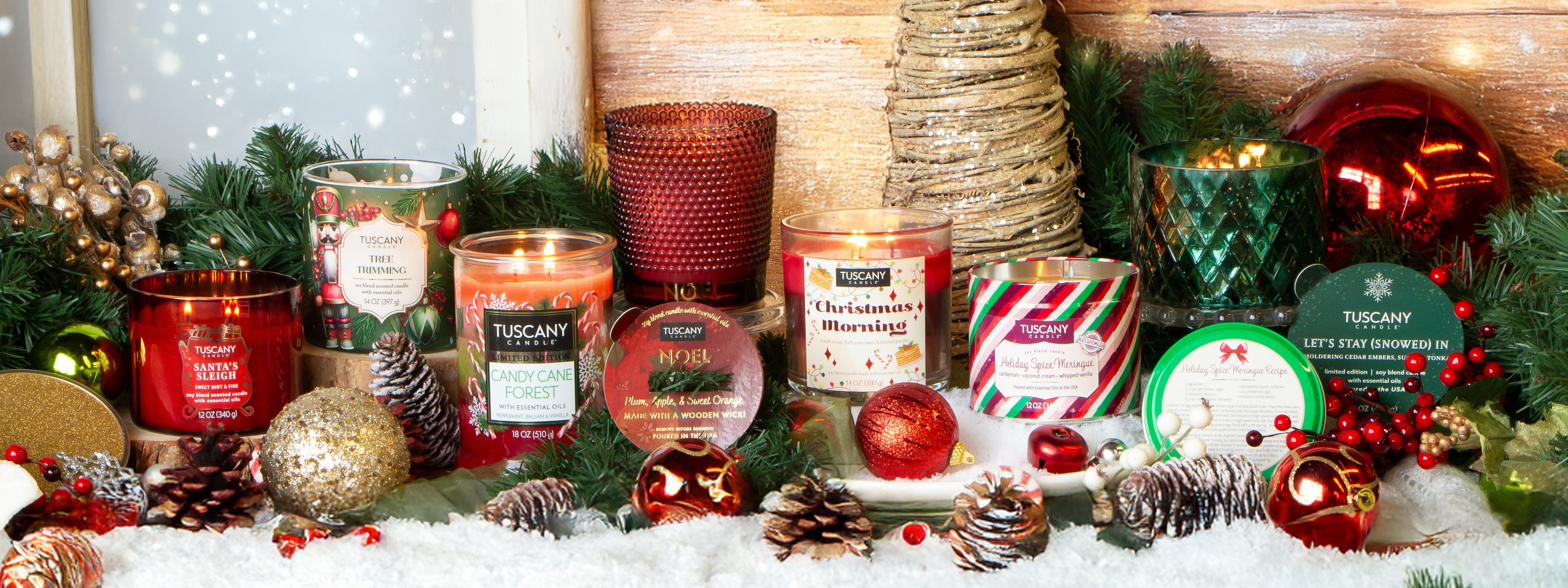 A variety of festive candles in glass jars and tins are displayed on a snow-like surface with pinecones, ornaments, and holiday greenery in the background.
