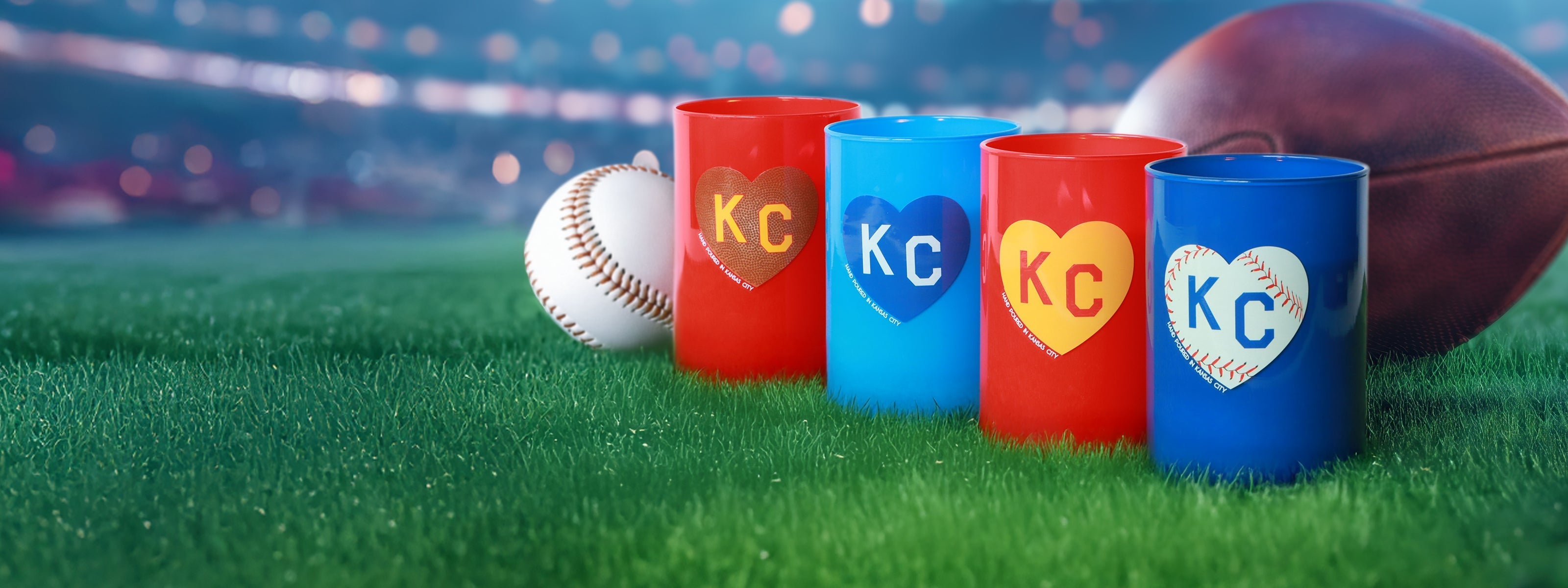 Four plastic cups, two red and two blue, decorated with "KC" inside heart shapes, are on grass with a baseball and football in the background under stadium lights.
