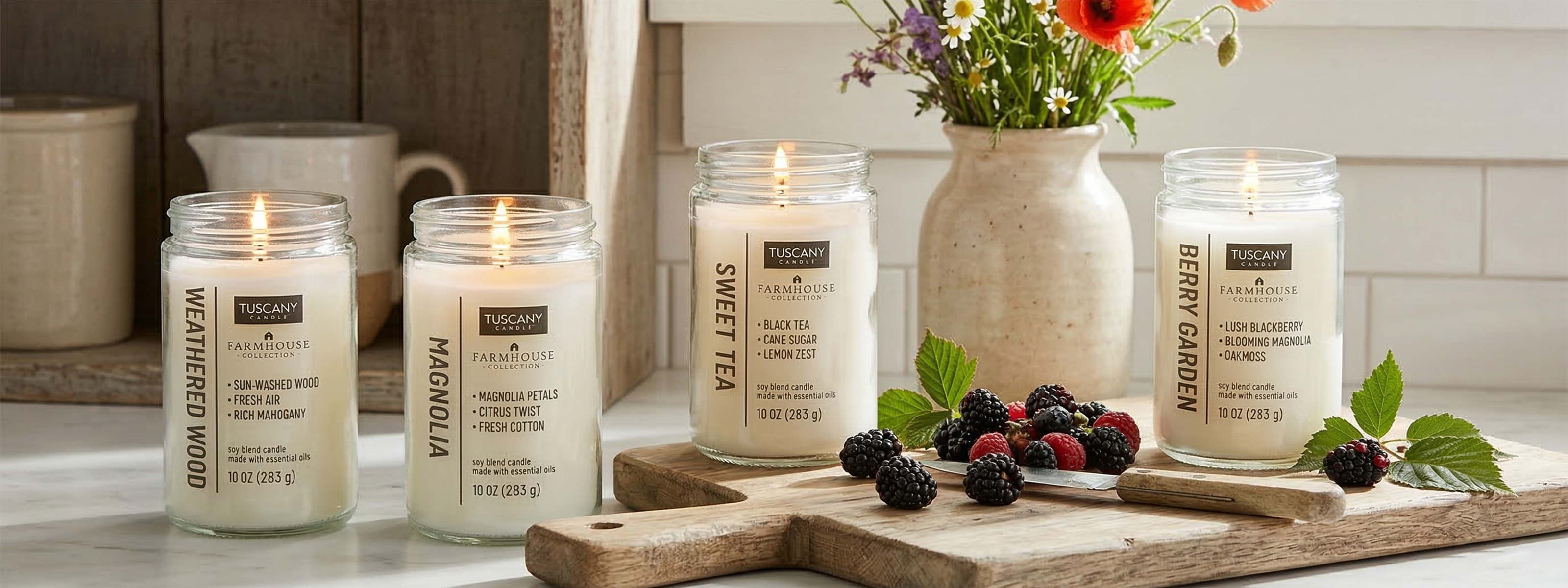 Four scented candles in glass jars are displayed on a kitchen counter next to a cutting board with berries and a vase of flowers.