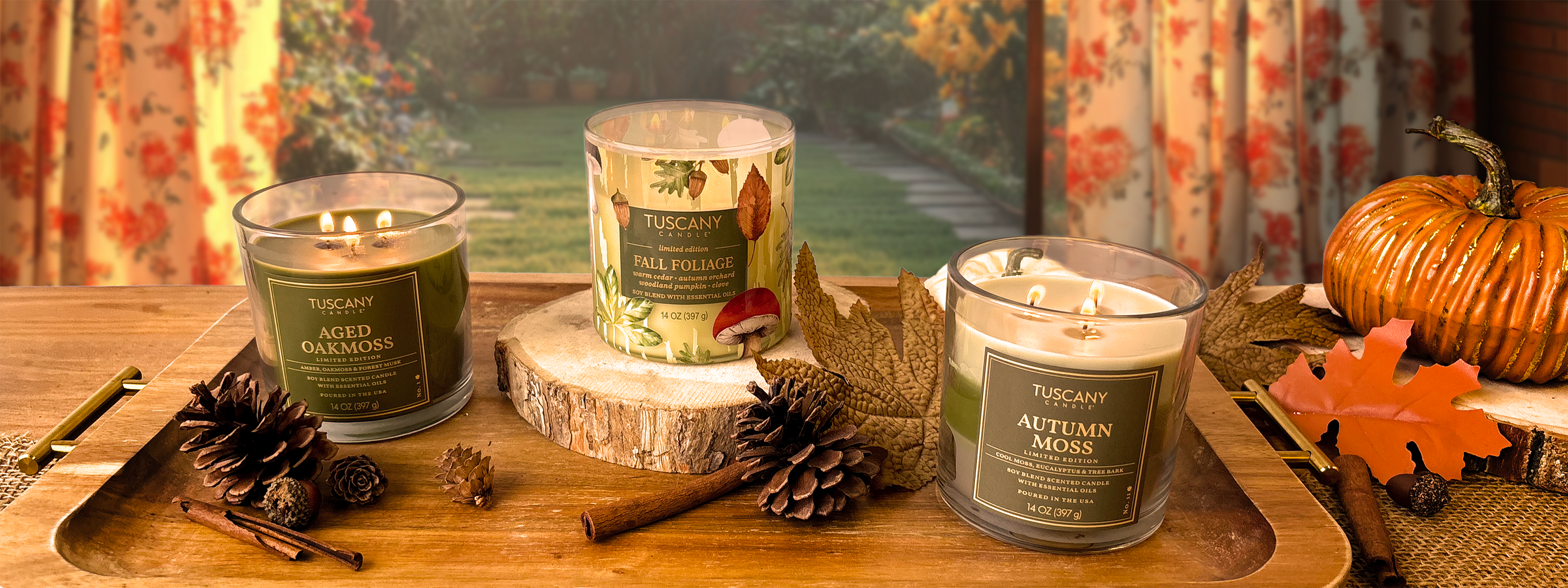 Three Tuscany brand candles labeled Aged Oakmoss, Fall Foliage, and Autumn Moss are displayed on a wooden tray with pinecones, leaves, and a pumpkin in a cozy fall setting.