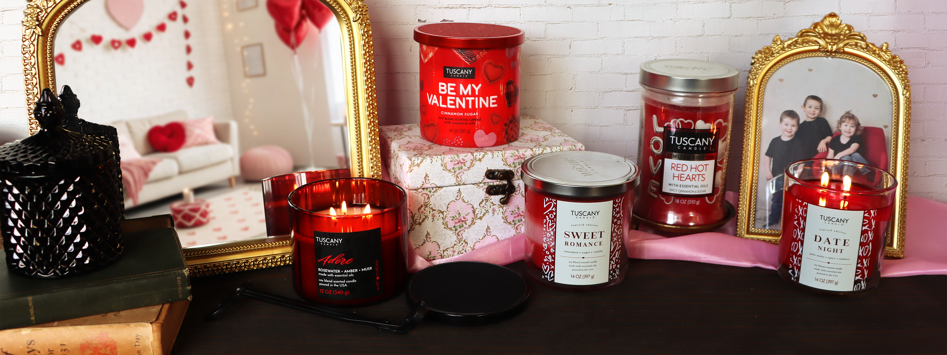 A variety of scented candles and jars are displayed on a table with framed family photos, a candle snuffer, books, and Valentine’s Day decorations in the background.
