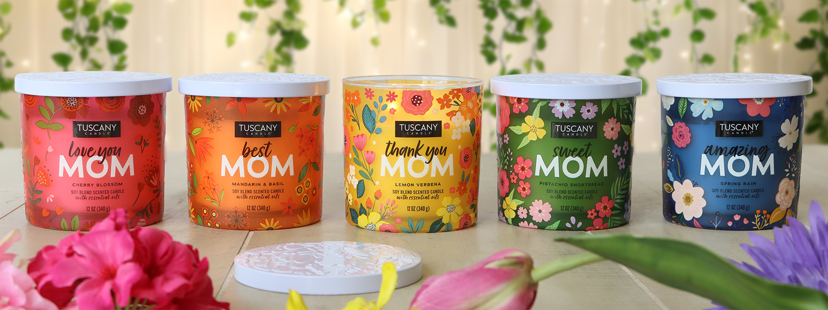 Five scented candles with floral designs and text: "Love you MOM," "Best MOM," "Thank you MOM," "Sweet MOM," "Amazing MOM." Various flowers are placed around them.