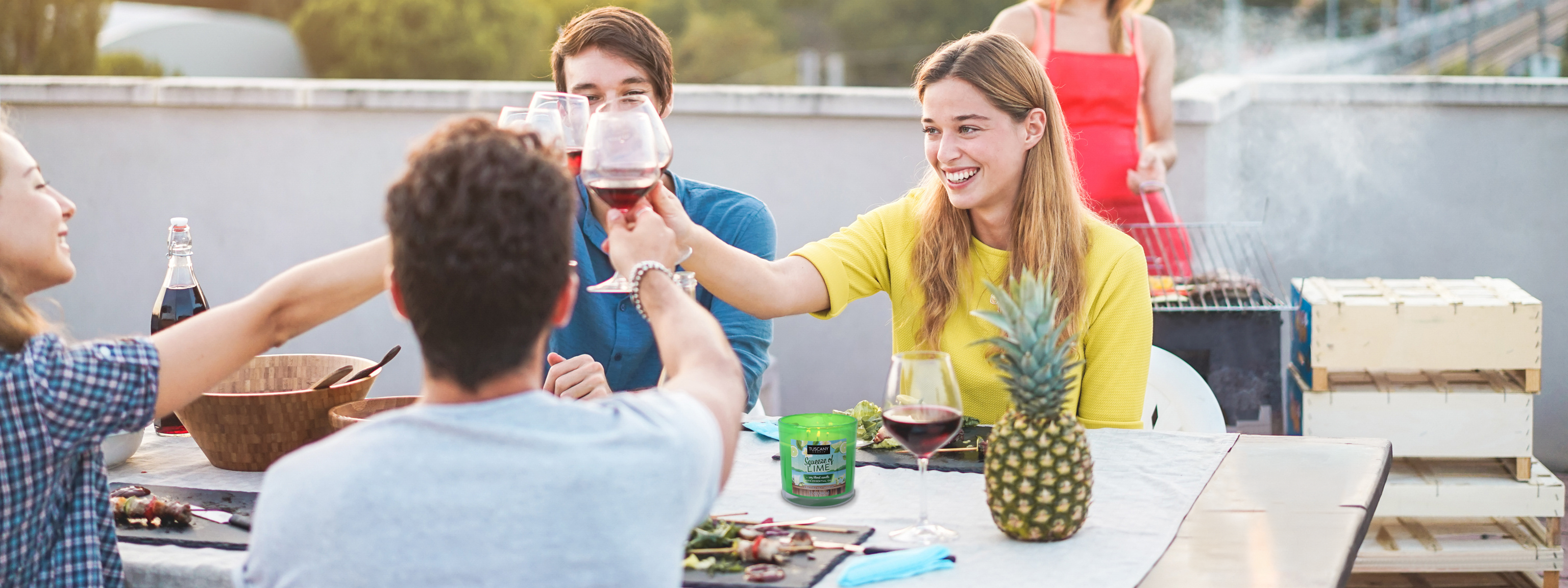 Four people sit around a table outdoors, toasting with wine glasses. A woman stands in the background tending to a grill. A pineapple and a green cup are on the table.
.
