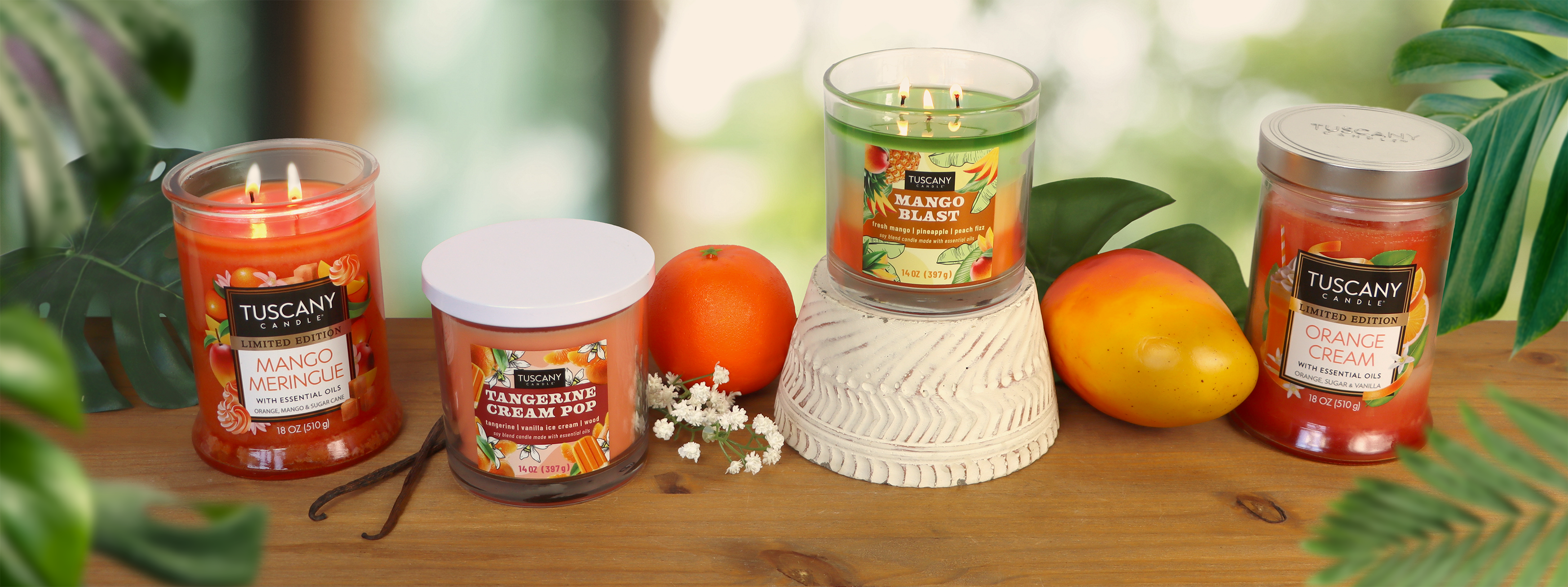 Four Tuscany Candle jars with citrus and mango scents are displayed on a wooden surface with oranges, a mango, and green leaves in the background.