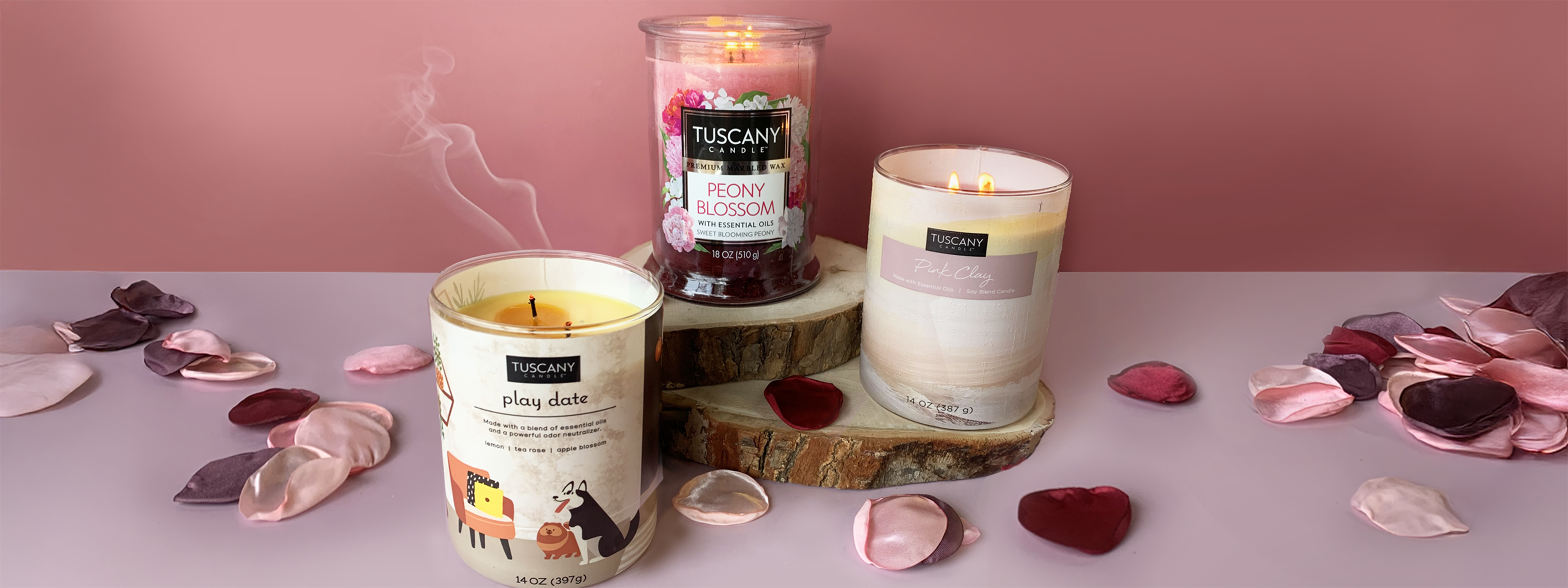 Three scented candles burning on wooden slabs, surrounded by rose petals. Candles labeled "Play date," "Peony Blossom," and "Pink Champagne" by Tuscany Candle.