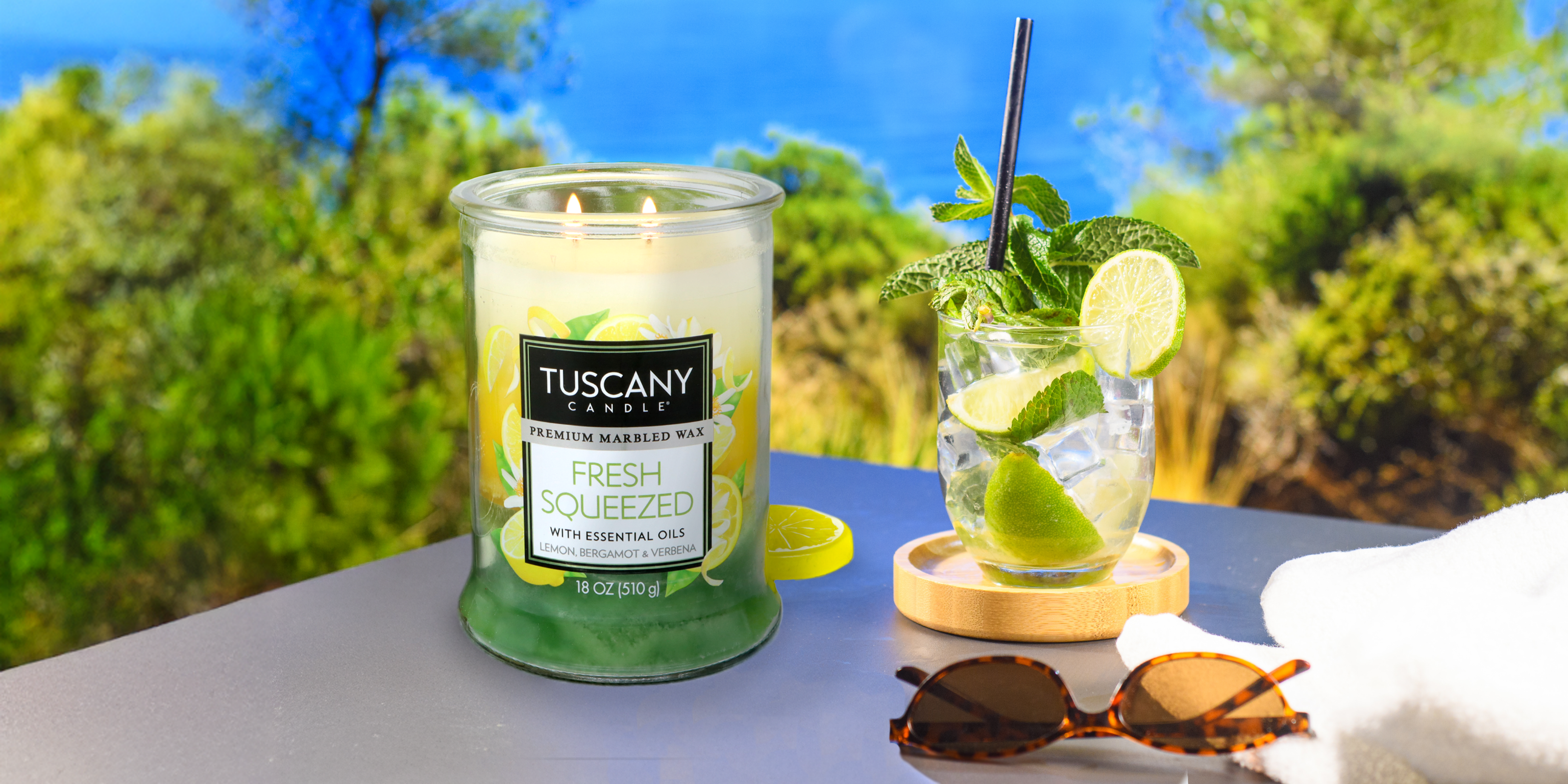 A Tuscany Candle labeled "Fresh Squeezed" sits beside a lime and mint cocktail, sunglasses, and a towel on a table with a blurred outdoor background of green trees and bright blue sky.