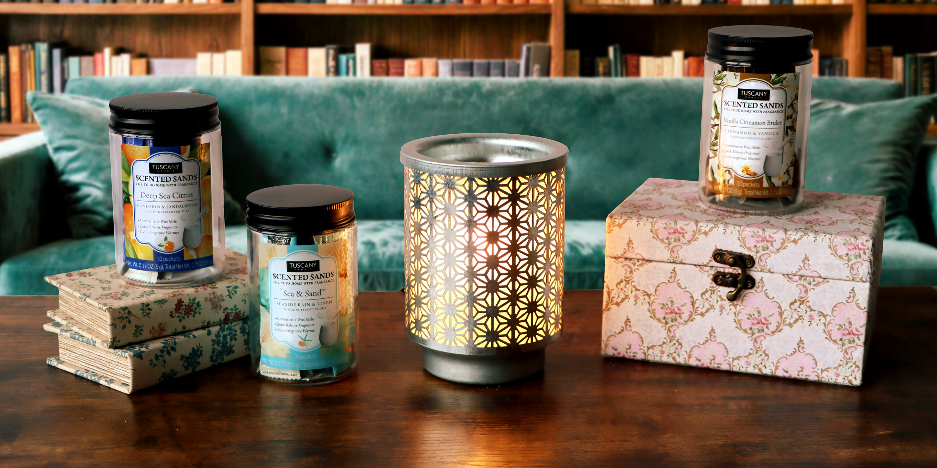 A decorative candle holder is lit on a wooden table, surrounded by three jars of scented sands, two floral-patterned boxes, and stacked books, with a green sofa and bookshelves in the background.