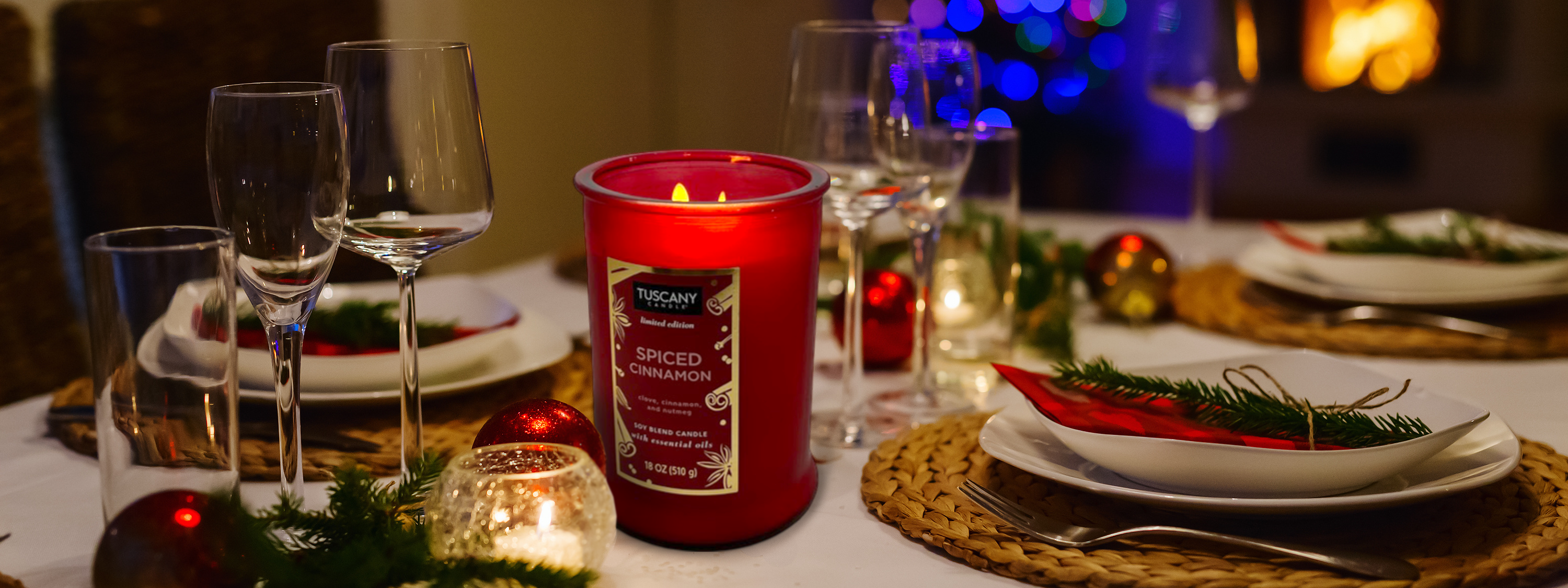 A festive table setting with a lit red candle labeled "Spiced Cinnamon," surrounded by wine glasses, plates, and Christmas decorations. In the background, a blurred Christmas tree is visible.