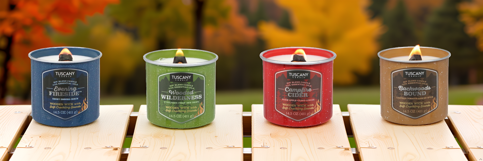 Four Tuscany brand candles—Evening Fireside, Woodland Wilderness, Campfire Cider, and Backwoods Bound—are lit and placed on a wooden table outdoors with autumn foliage in the background.