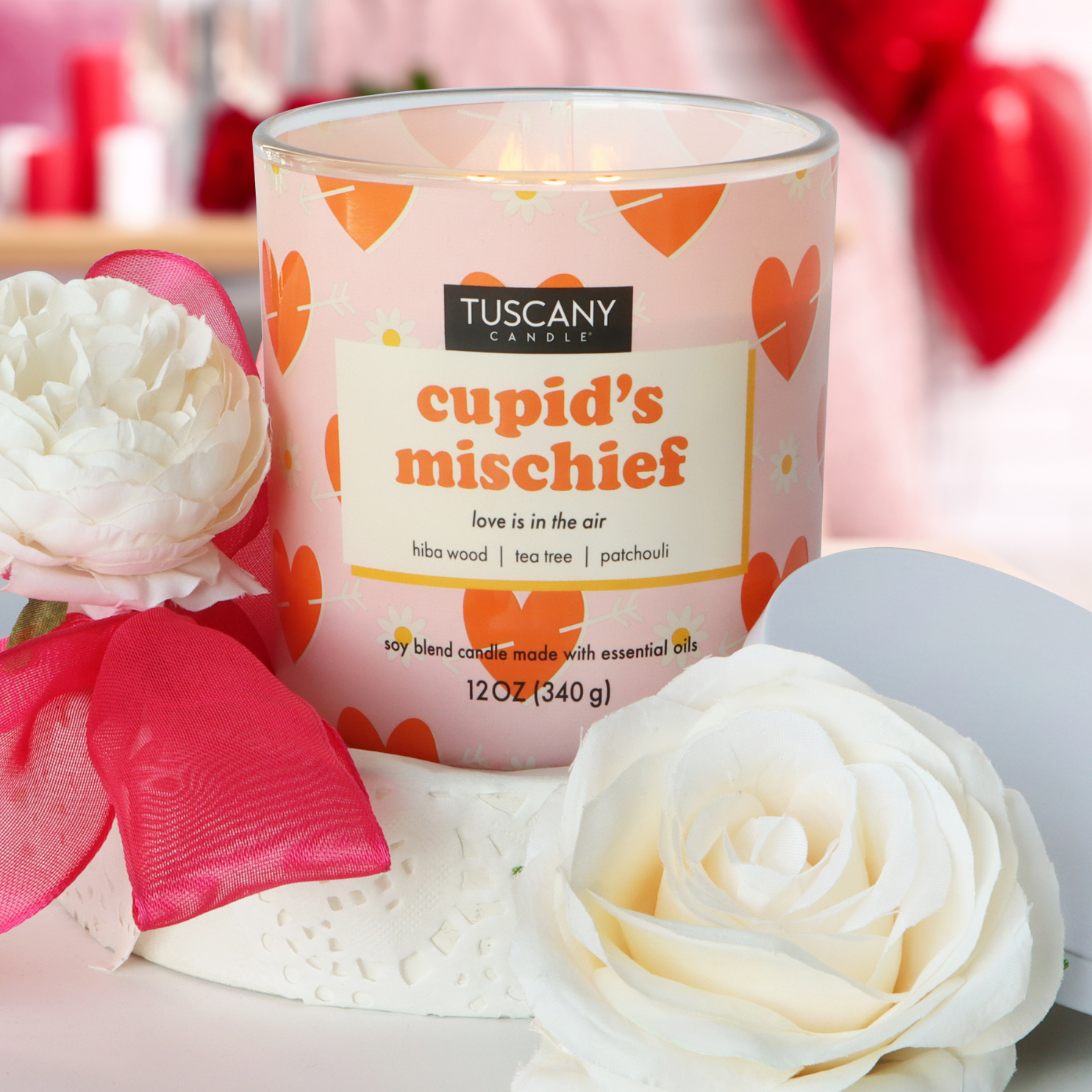 A romantic scented candle named "Cupid's Mischief: Patchouli Scented Valentine's Day Candle" by Tuscany Candle® SEASONAL, featuring notes of hiba wood, tea tree, and patchouli, is elegantly displayed among white flowers and a red ribbon on a lace doily. Ideal for setting the mood on Valentine's Day.