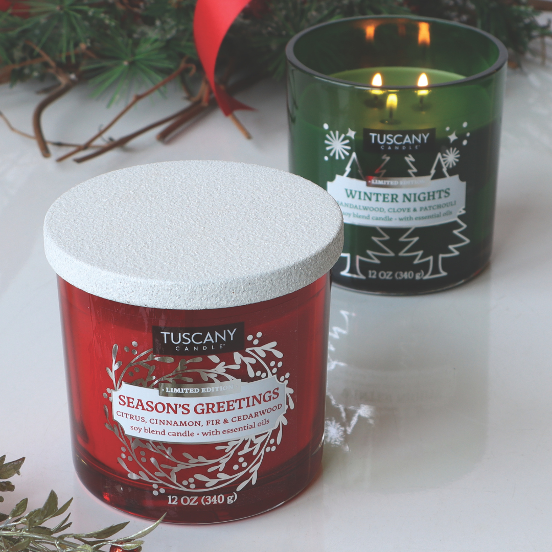 A red Tuscany Candle® SEASONAL "Season's Greetings Scented Jar Candle" (12 oz) and a green one labeled "Winter Nights" are placed on a table surrounded by holiday decorations in the background.