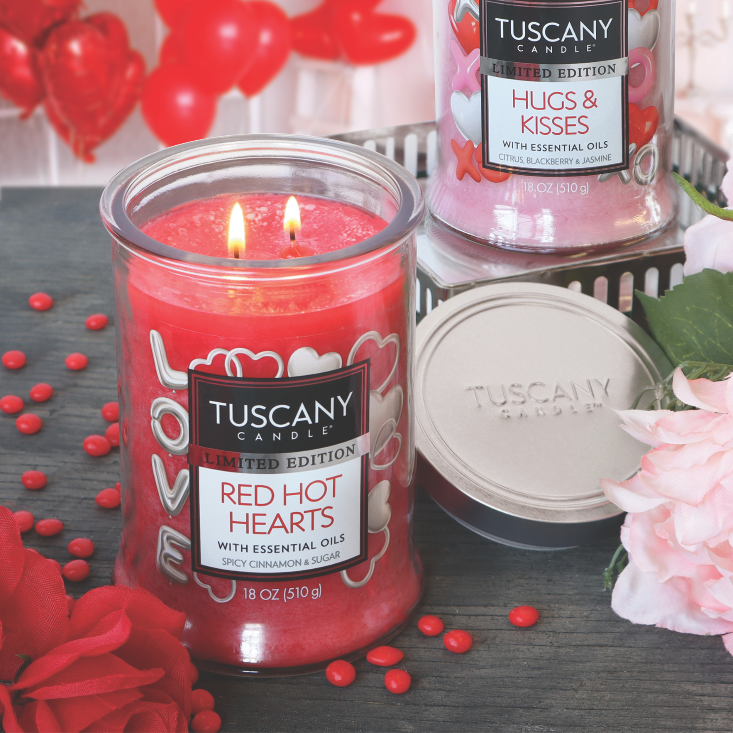 The "Red Hot Hearts" cinnamon-scented Valentine's Day candle, offered by Tuscany Candle® SEASONAL in an 18 oz size, makes the perfect romantic addition when paired with a second red candle on a wooden surface accented by flowers and a silver lid.