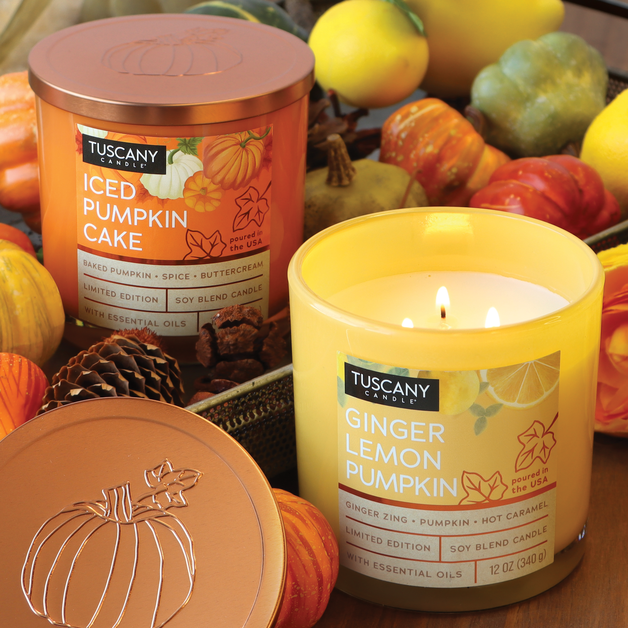 Two Tuscany Candle® SEASONAL candles from the Copper Harvest Collection: one labeled "Iced Pumpkin Cake Long-Lasting Scented Jar Candle (12 oz)," the other burning and labeled "Ginger Lemon Pumpkin." Both are surrounded by small decorative pumpkins and gourds, filling the room with a delightful autumn fragrance.