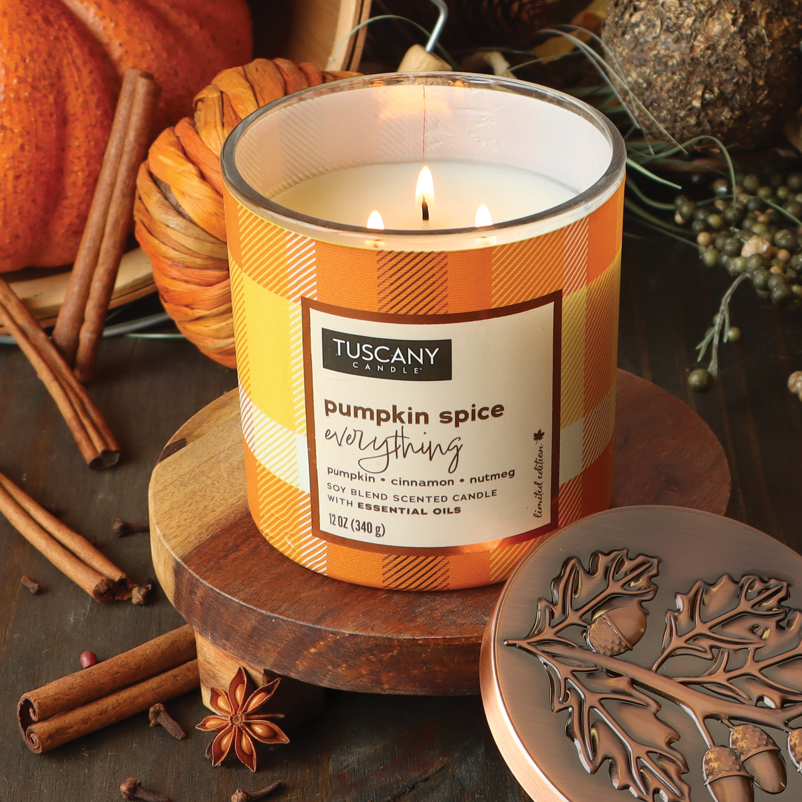 A lit "Pumpkin Spice Everything" Long-Lasting Scented Jar Candle (12 oz) from the Tuscany Candle® SEASONAL Autumn Flannels Collection sits in a plaid container on a wooden surface near cinnamon sticks, pine cones, a pumpkin, and a decorative lid.