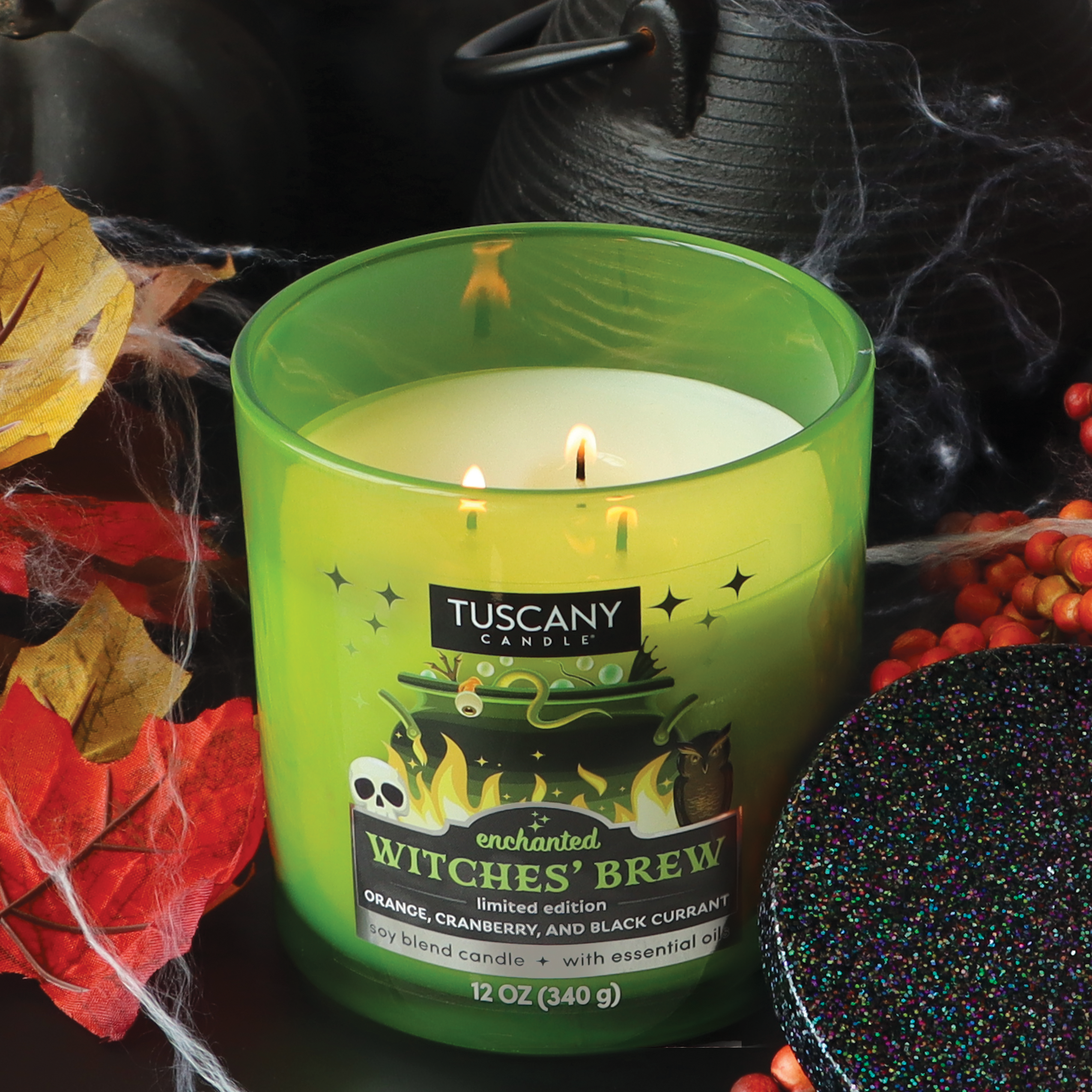 A lit "Enchanted Witches Brew Long-Lasting Scented Jar Candle (12 oz)" in a green jar sits surrounded by colorful autumn leaves and berries. The candle, branded "Tuscany Candle® SEASONAL," has a black glittery lid beside it, perfect for adding an enchanting touch to Halloween gatherings with its mystical aroma.