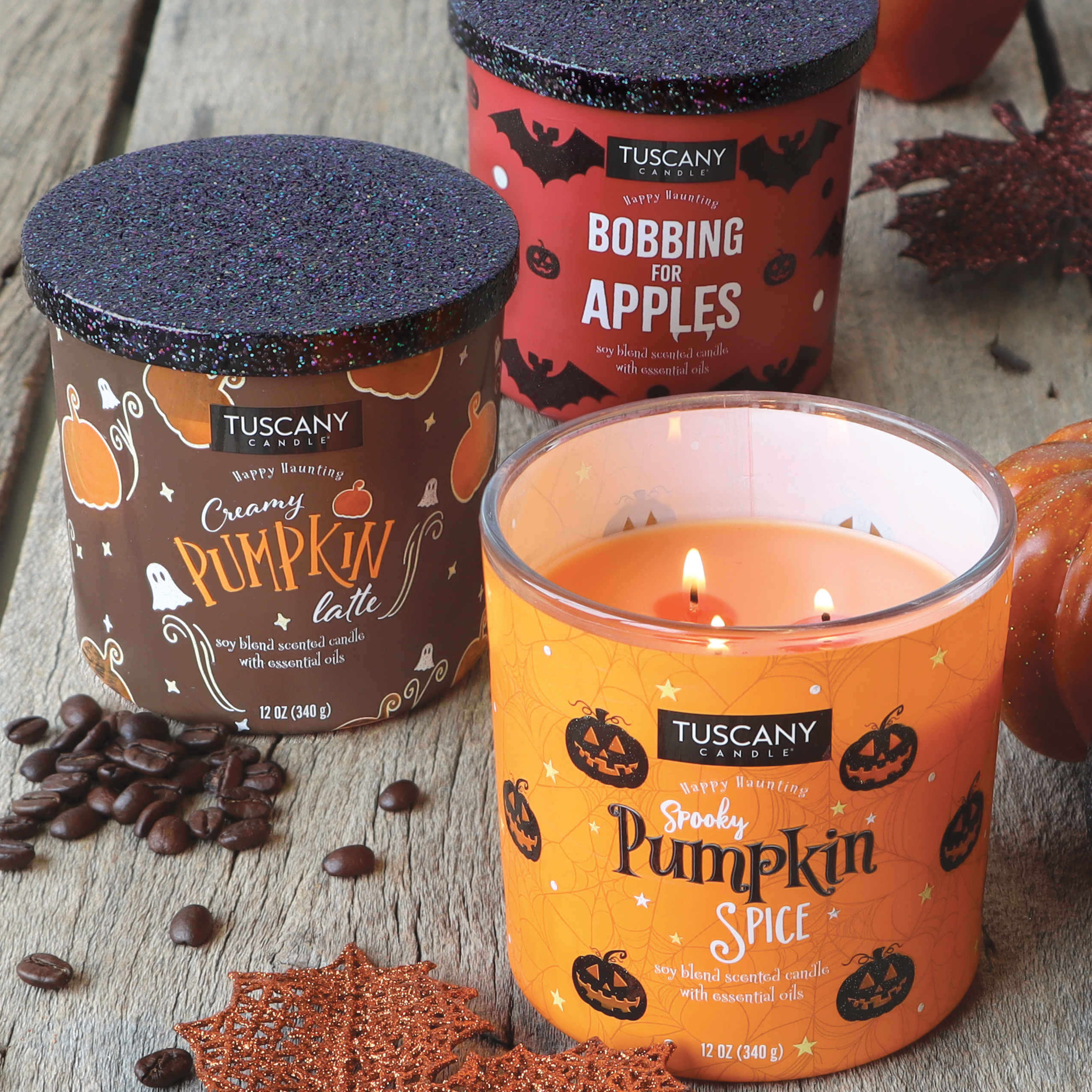 Three Tuscany Candle® SEASONAL candles are displayed on a wooden surface: "Creamy Pumpkin Latte," "Bobbing For Apples Long-Lasting Scented Jar Candle (12 oz)," and the lit "Spooky Pumpkin Spice." Each candle offers an inviting autumn scent that perfectly captures the essence of the season.