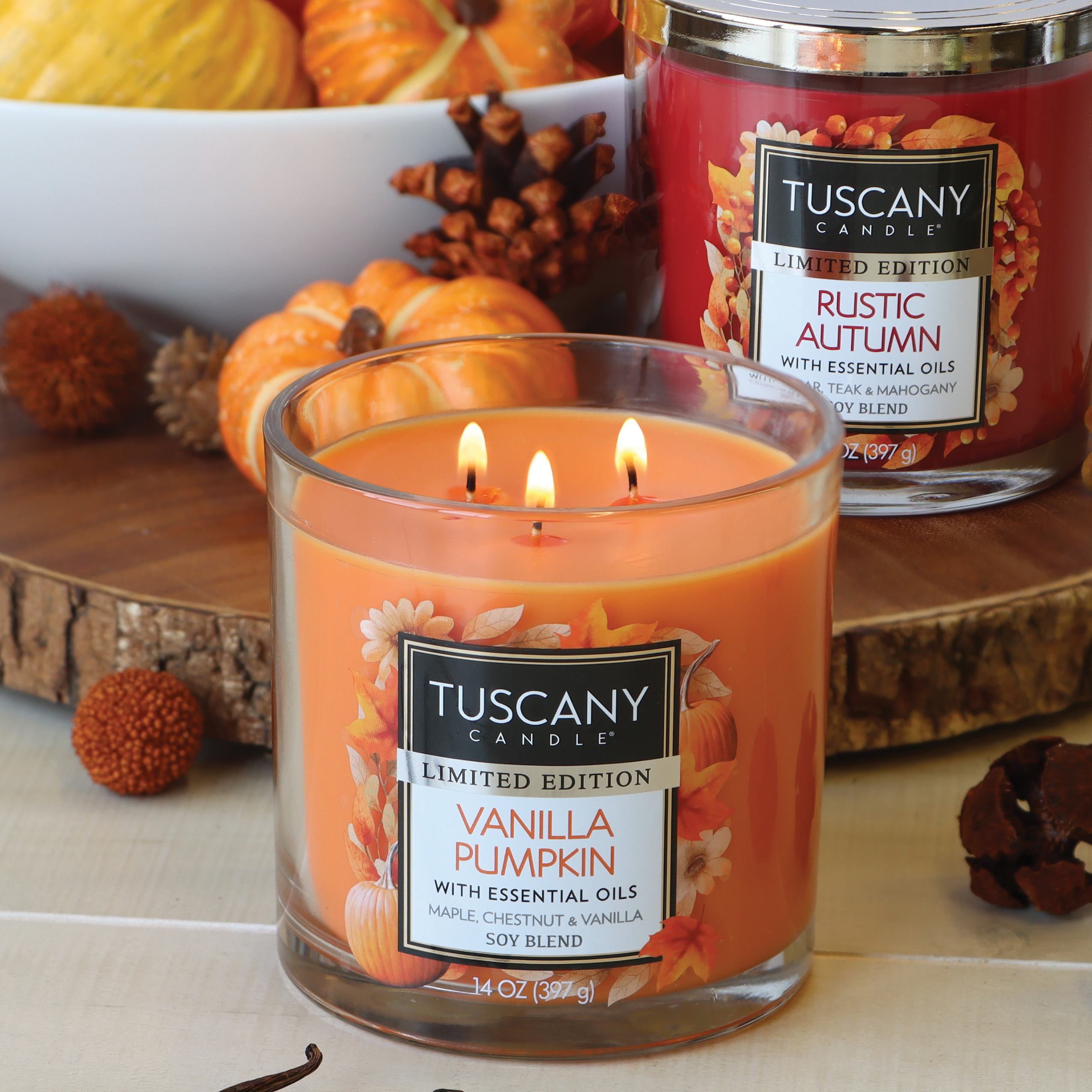 Two fragrant candles from the Fall Collection, one labeled "Vanilla Pumpkin Long-Lasting Scented Jar Candle (14 oz)" and the other "Rustic Autumn," both by Tuscany Candle® SEASONAL, are lit on a wooden surface with decorative pumpkins and autumn elements in the background.