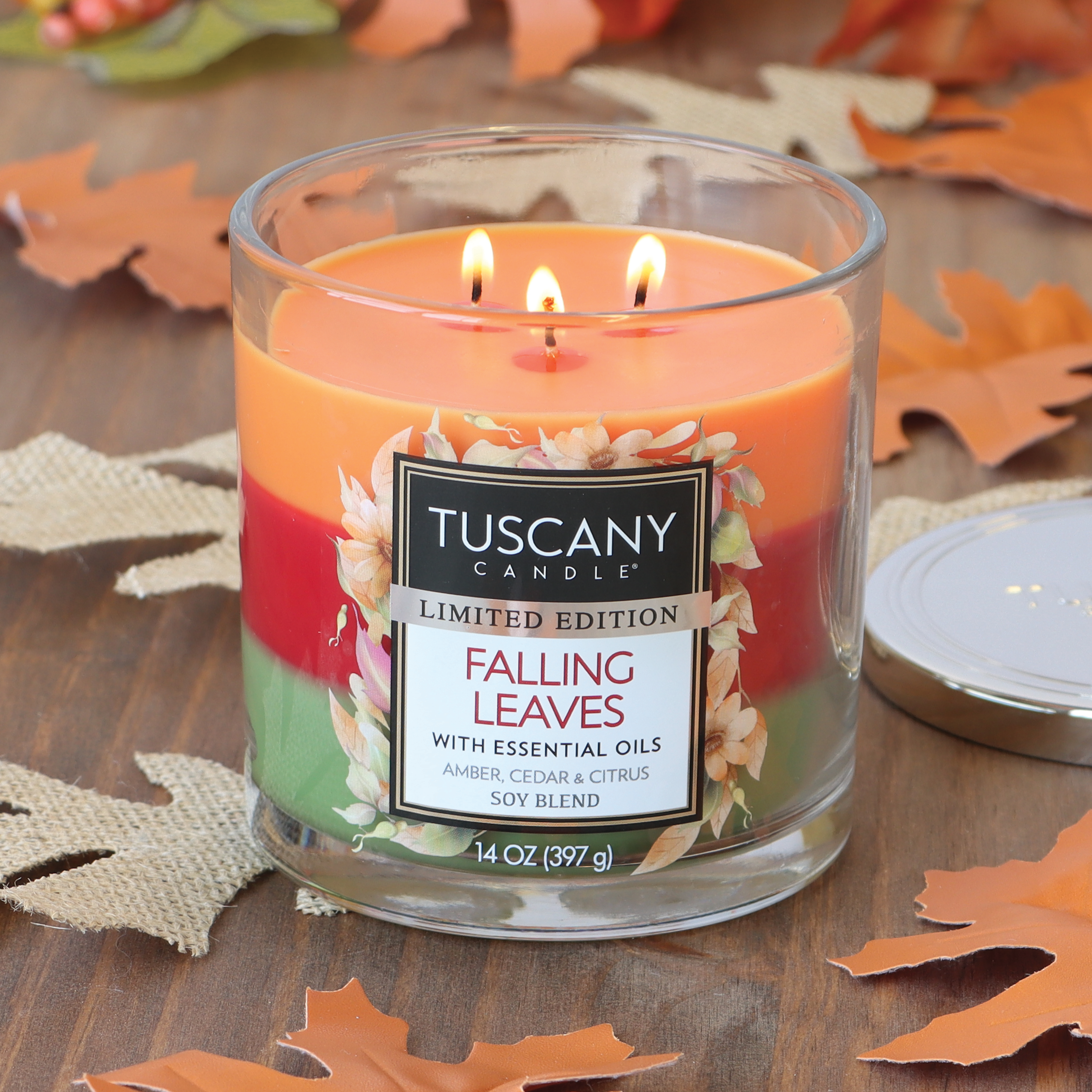 A lit Falling Leaves Long-Lasting Scented Jar Candle (14 oz) by Tuscany Candle® SEASONAL, featuring essential oils with woodland amber, cedar, and citrus scents. The candle boasts a layered design and is surrounded by decorative autumn leaves, perfectly capturing the essence of the season's fragrance.