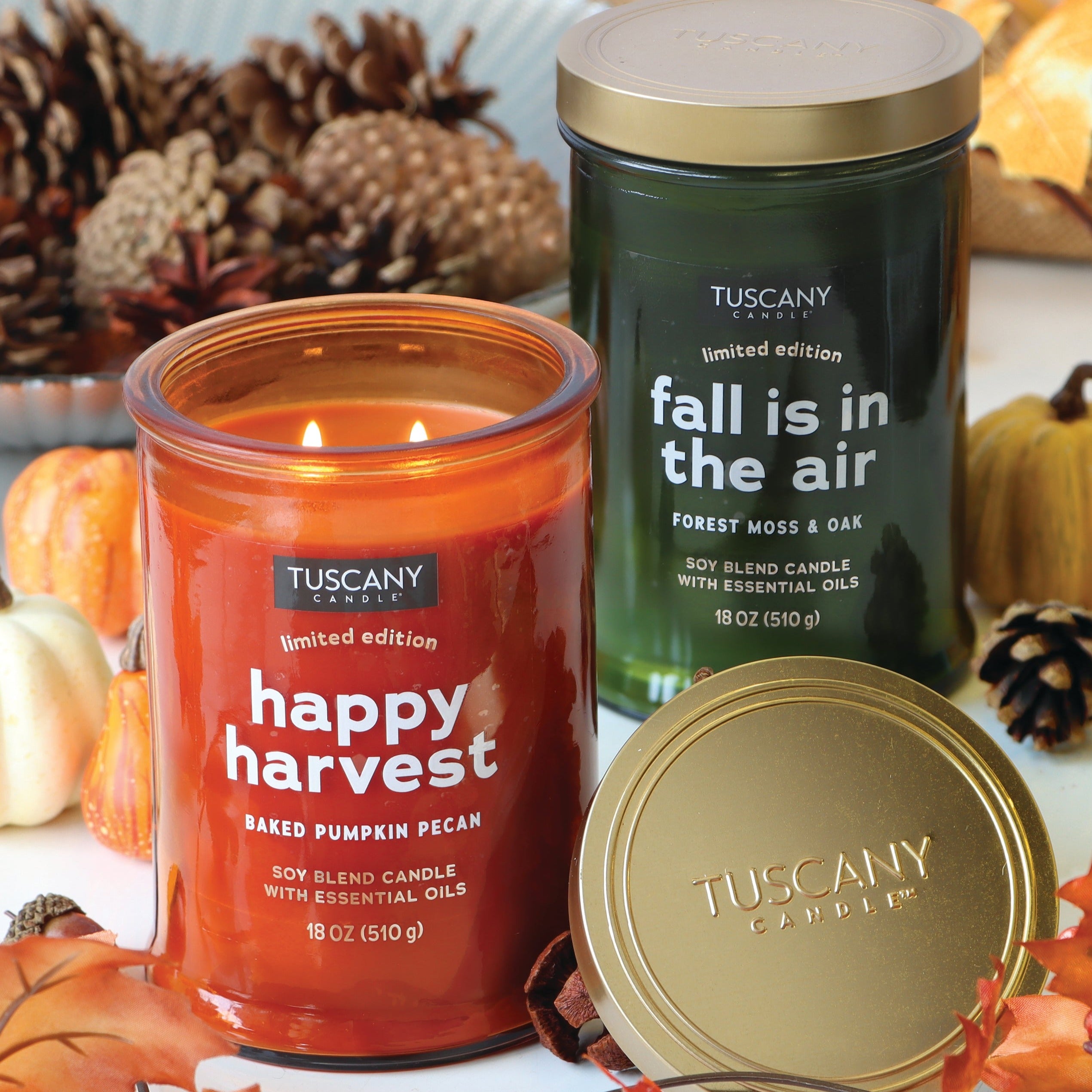 Two Tuscany Candle® SEASONAL candles from the Gather Together Collection — one, the "Happy Harvest Long-Lasting Scented Jar Candle" in orange and the other labeled "Fall is in the Air" in green — emit a delightful fall fragrance. They are surrounded by autumn leaves, mini pumpkins, and pine cones.