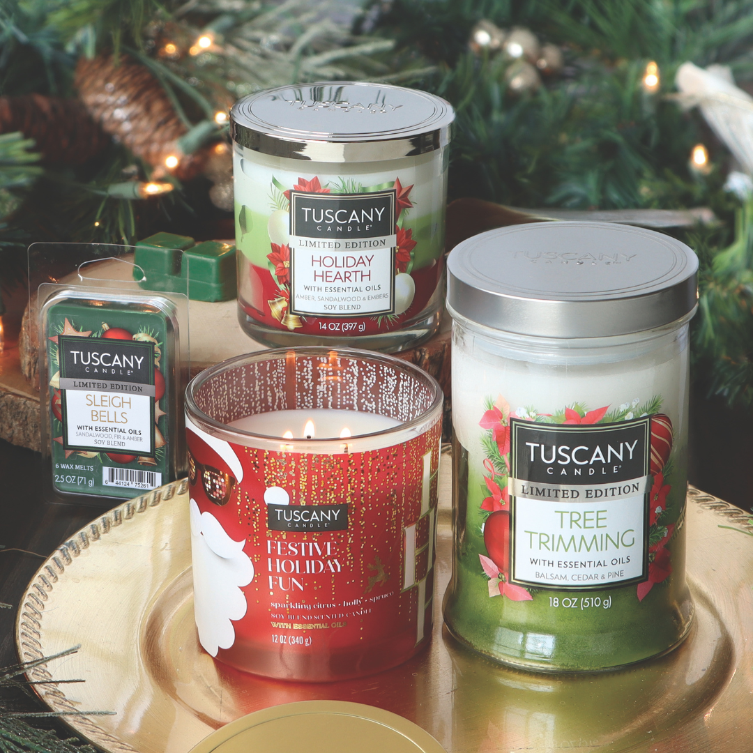 A selection of Tuscany Candle® SEASONAL products, including the Holiday Hearth Long-Lasting Scented Jar Candle (14 oz), arranged on a gold tray with aromatic options such as Festive Holiday Fun, Tree Trimming, and Sleigh Bells, accented by lush greenery.