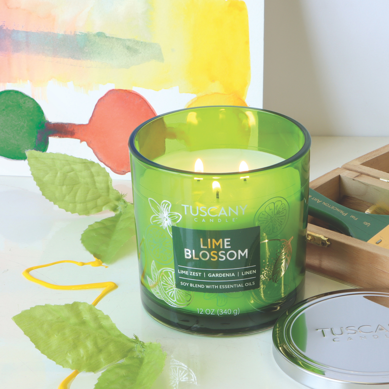 A Lime Blossom: Linen Scented Jar Candle from Tuscany Candle® EVD's Painters' Palette Collection flickers on the table, surrounded by gardenia petals and art supplies, with a colorful painting in the background.