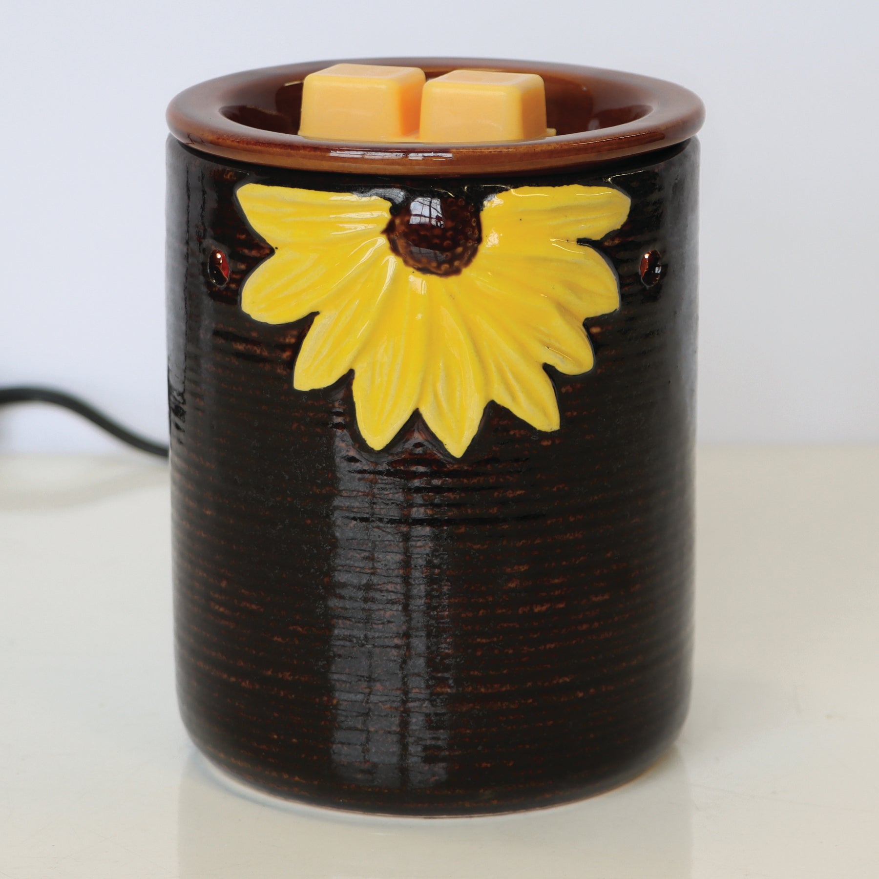 A dark brown Sunflower Wax Melt Warmer with a yellow flower design, holding two yellow Tuscany Candle® SEASONAL wax melts, is placed on a white surface.