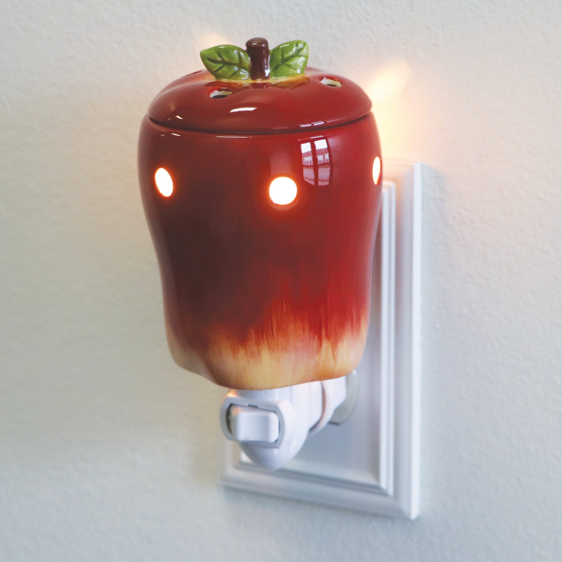 An Apple Outlet Wax Melt Warmer from Tuscany Candle® SEASONAL, glowing with small lights from a low wattage light bulb, plugs into a wall outlet for a cozy ambiance.