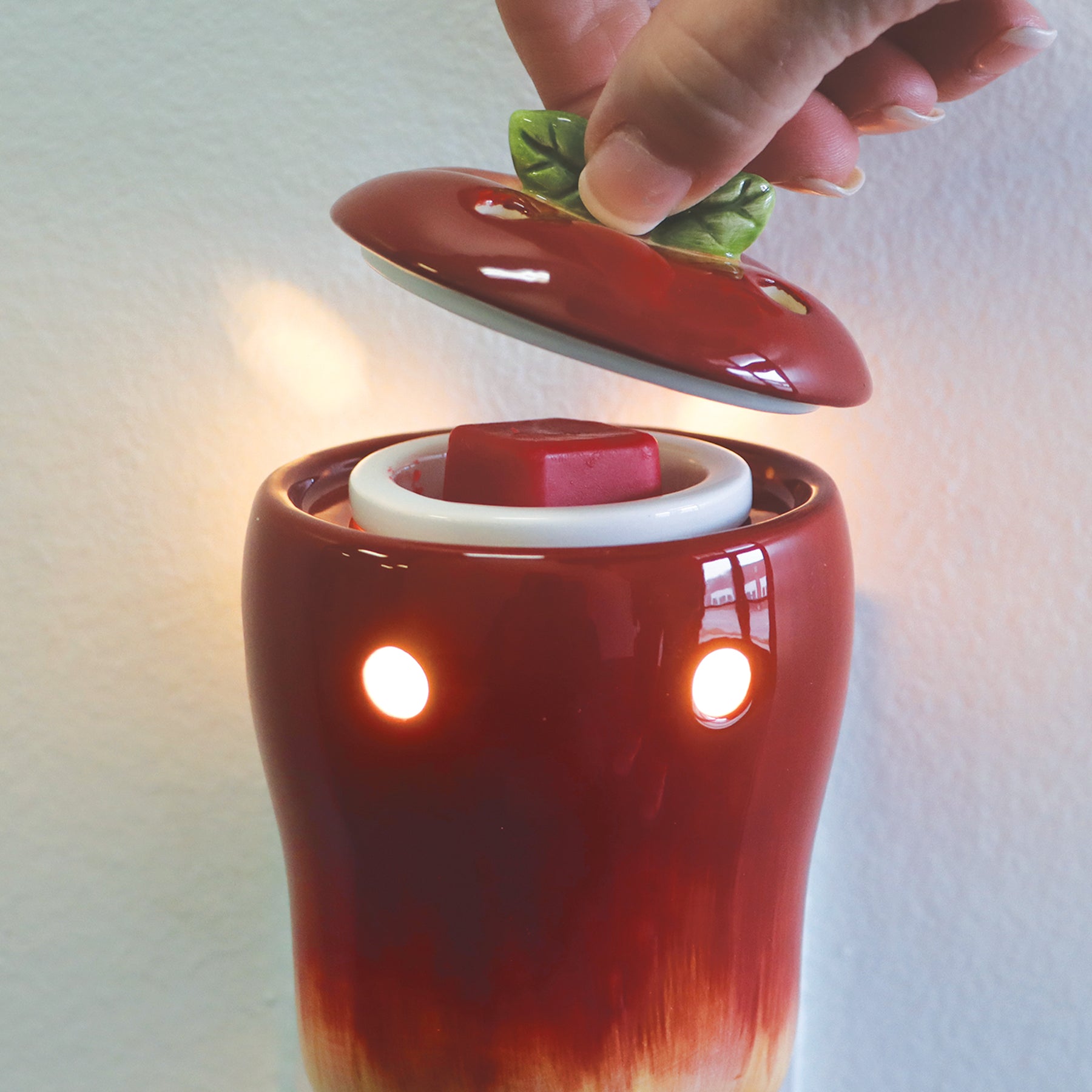 A hand lifts the lid of a red ceramic warmer, resembling an Apple Outlet Wax Melt Warmer, revealing a red Tuscany Candle® SEASONAL wax melt inside, with two lit holes emitting light from its front.