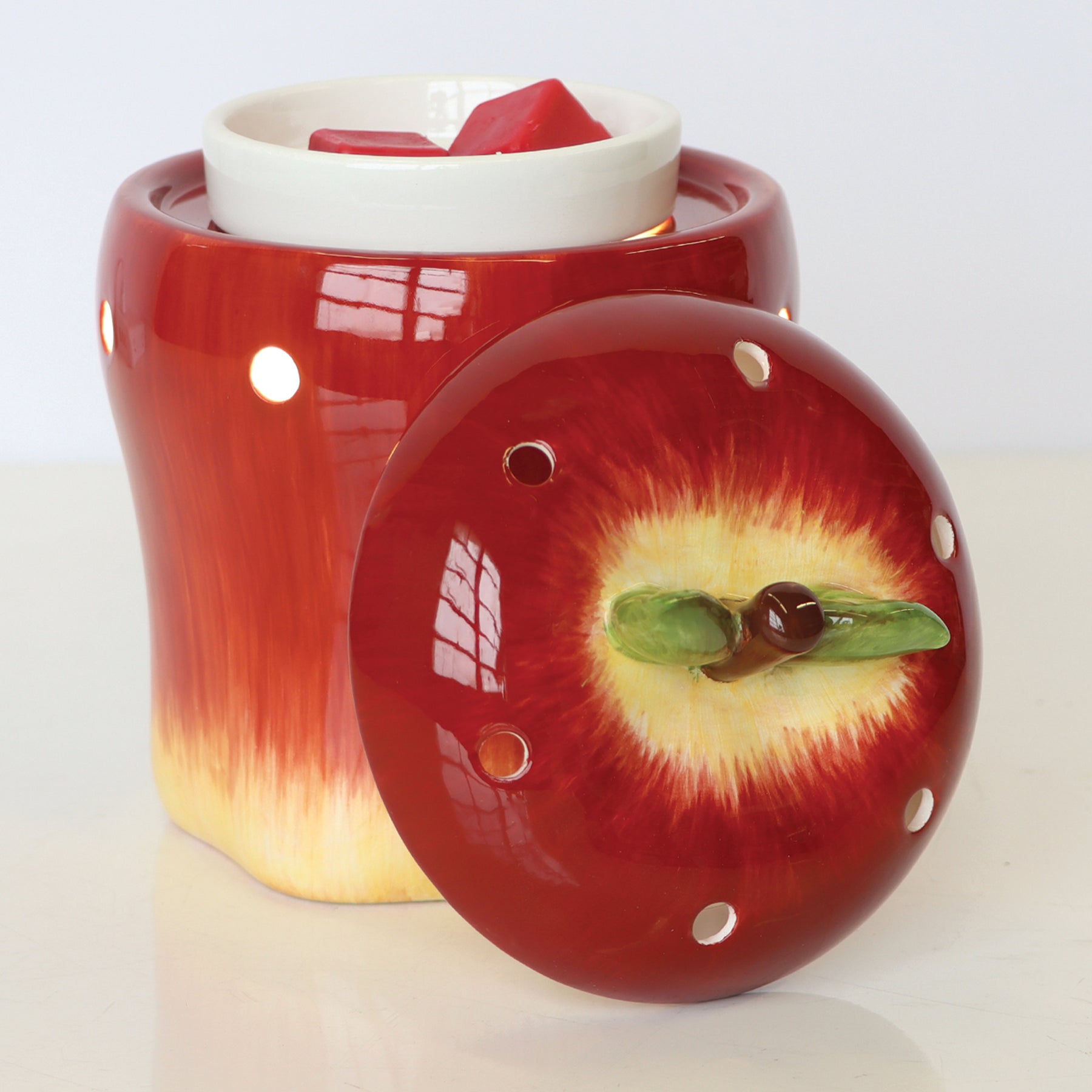 An Apple Wax Melt Warmer by Tuscany Candle® SEASONAL, with a removable lid, containing Tuscany Candle wax melts inside, is placed on a white surface.