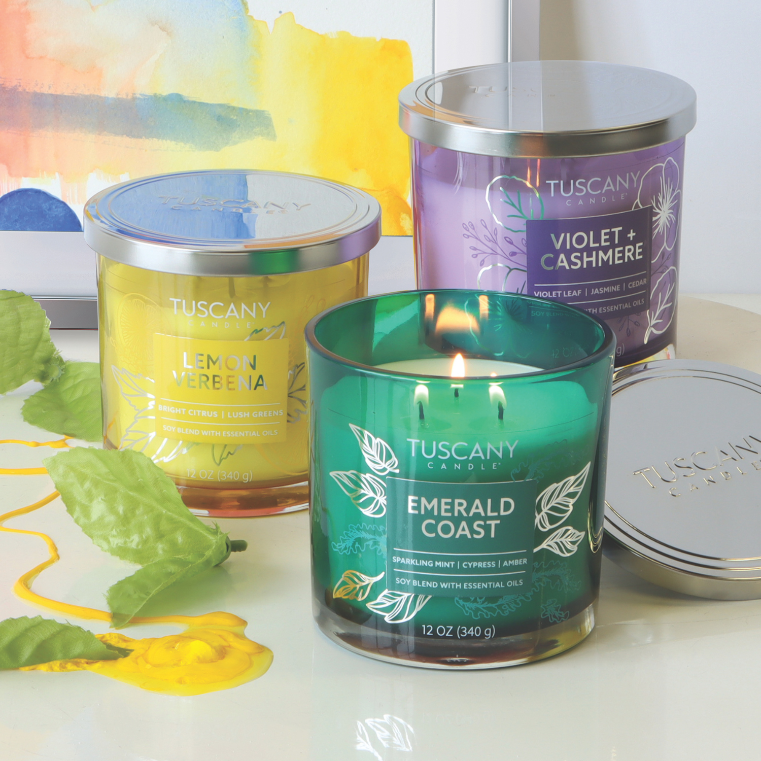 Three Lemon Verbena, Violet + Cashmere, and Emerald Coast scented candles are displayed against a colorful background. The Violet + Cashmere candle by Tuscany Candle® EVD fills the air with an alluring jasmine fragrance enriched by subtle cedar notes for a deep aromatic experience.