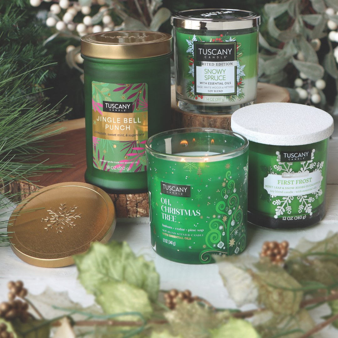 A set of four Snowy Spruce Long-Lasting Scented Jar Candles by Tuscany Candle® SEASONAL, each with festive labels, arranged on a wooden surface and surrounded by pine branches and white flowers.