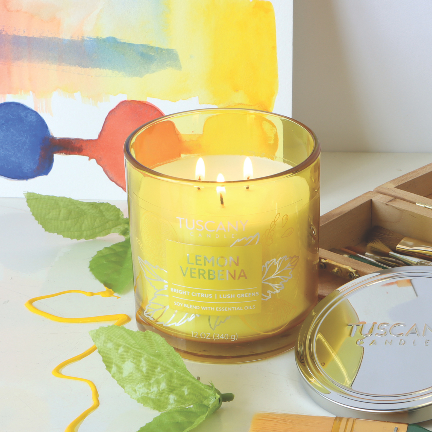 A lit Lemon Verbena: Meyer Lemon Scented Jar Candle (12 oz) by Tuscany Candle® EVD from the Painters' Palette Collection sits on a table with green leaves, paintbrushes, and a colorful painting.