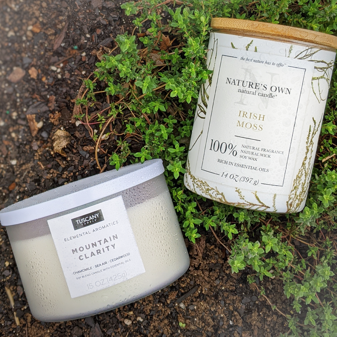 A Mountain Clarity scented jar candle (15 oz) from the Elemental Aromatics Collection by Tuscany Candle, and a tin sitting on the ground, creating a serene atmosphere of tranquility for enhanced well-being.