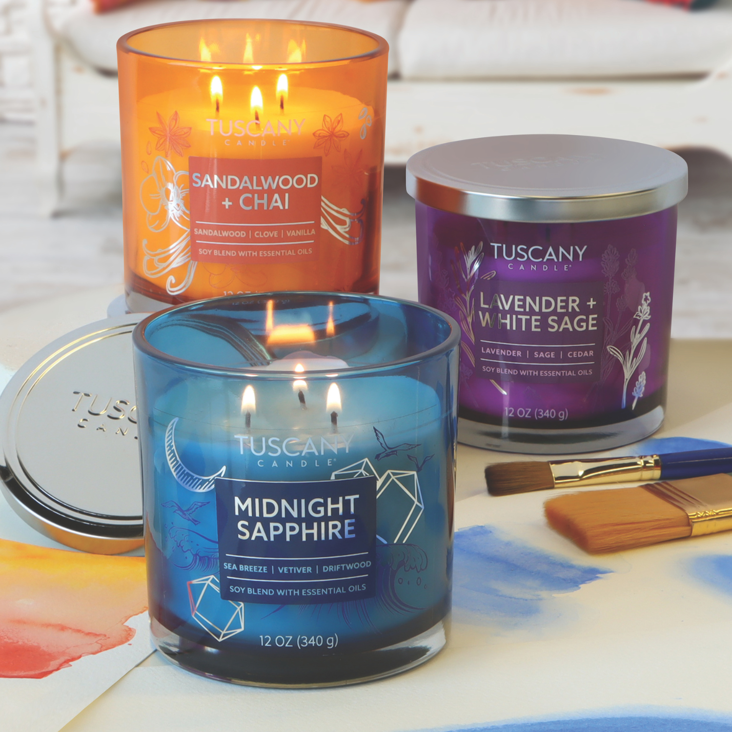 Three Tuscany Candle® EVD candles are displayed: Sandalwood + Chai, Lavender + White Sage, and Midnight Sapphire. The Sandalwood + Chai candle from the Painters' Palette Collection offers a warm vanilla bean aroma. A paintbrush and metallic lid are nearby.