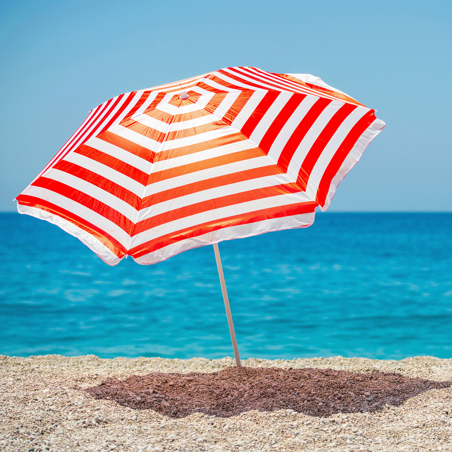 The Sea & Sand Long-Lasting Scented Jar Candle (18 oz) by Tuscany Candle® EVD evokes a beach-inspired atmosphere reminiscent of a red and white striped umbrella planted in the sand on a beach, with the ocean and a clear blue sky in the background.