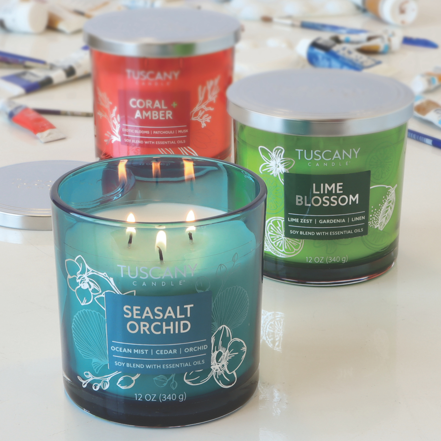 A 12 oz Seasalt Orchid: Ocean Scented Jar Candle from the Tuscany Candle® EVD Painters' Palette Collection sits on a table with art supplies, filling the room with a fragrance reminiscent of salty ocean mist.