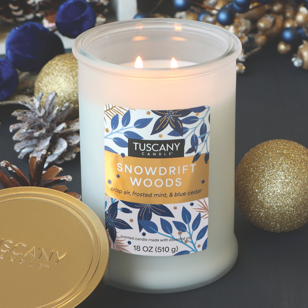 A lit Snowdrift Woods Scented Jar Candle (18 oz) by Tuscany Candle® SEASONAL is surrounded by gold and blue ornaments and pine cones.