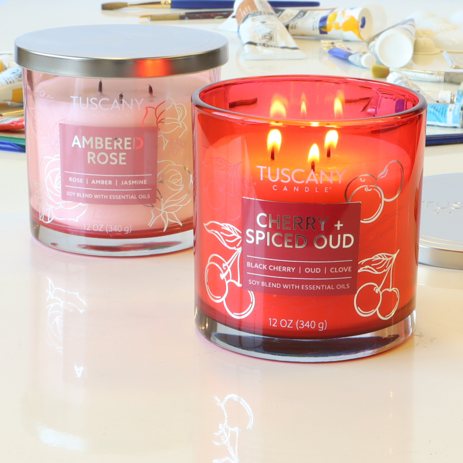 Two Tuscany candles on a table: one is a red glass labeled "Cherry + Spiced Oud," the other is an Ambered Rose: Jasmine Scented Jar Candle (12 oz) from the Painters' Palette Collection, evoking blooming roses. Assorted art supplies are visible in the background.