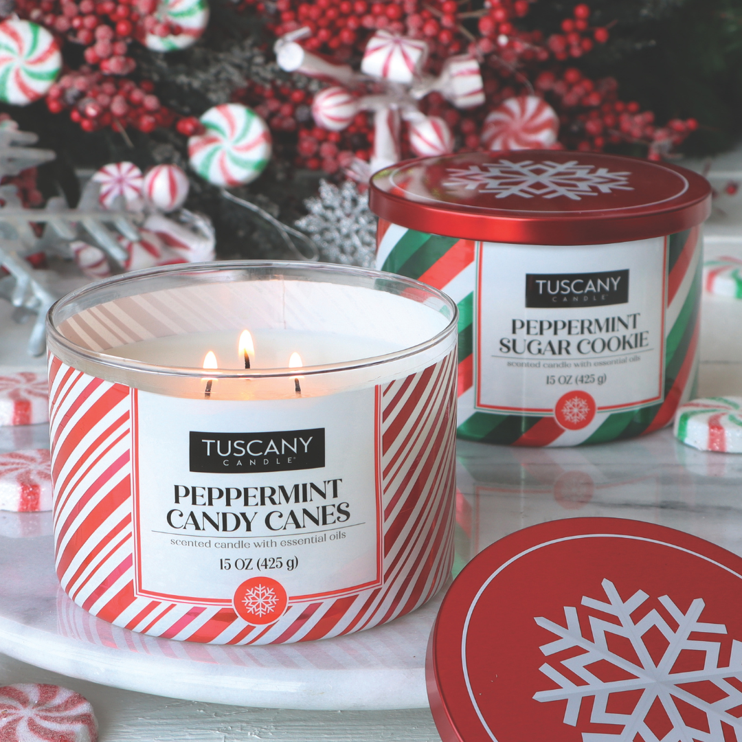 Two Tuscany Candle® SEASONAL scented jar candles on a table, one labeled "Peppermint Candy Canes Scented Jar Candle (15 oz)" and the other "Peppermint Sugar Cookie," surrounded by peppermint candies.