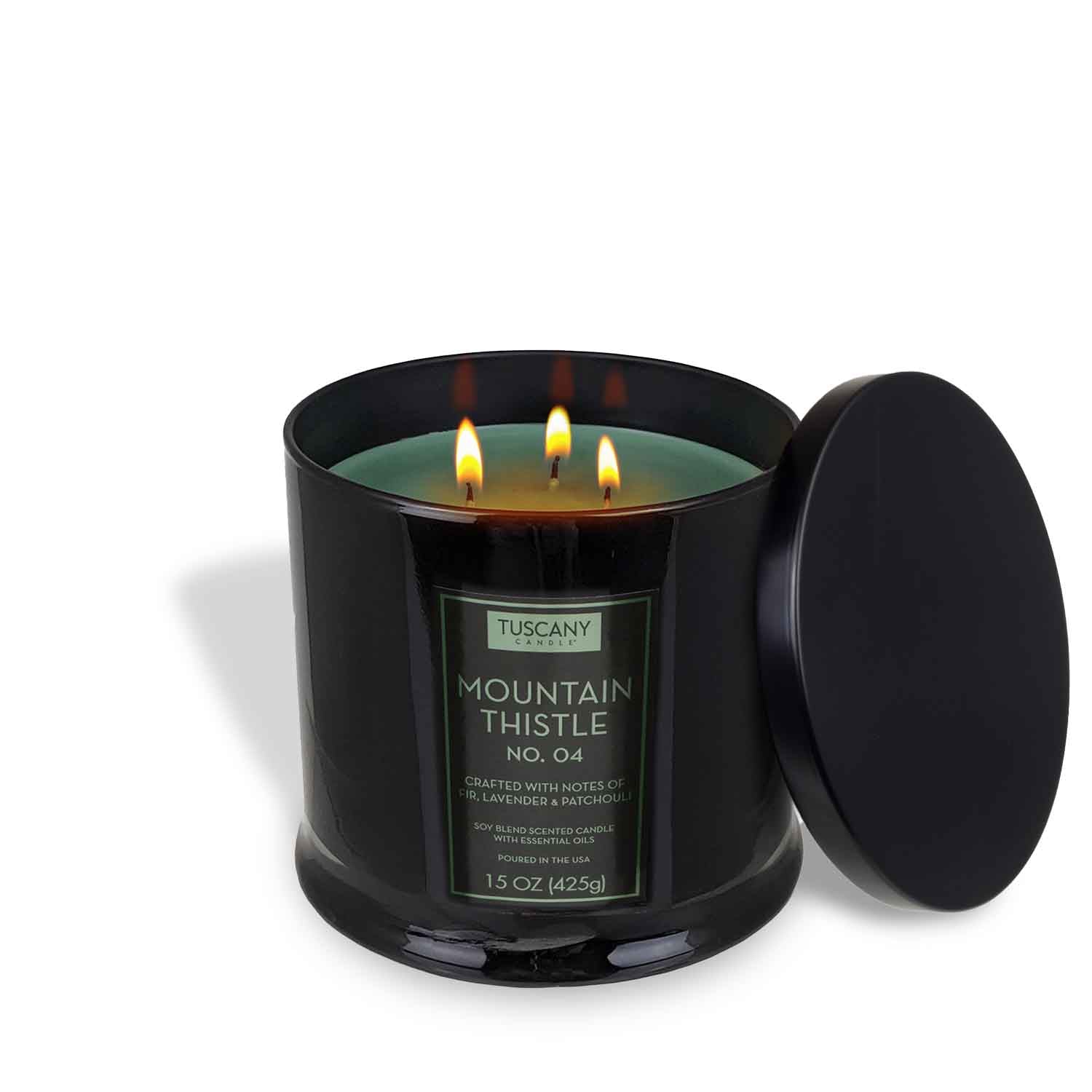 A black 15 oz Mountain Thistle Scented Jar Candle (No. 04) by Tuscany Candle® EVD, part of the Homme + Heritage Collection, with three wicks lit and its matching black lid beside it, fills the room with a subtle fir needle scent.