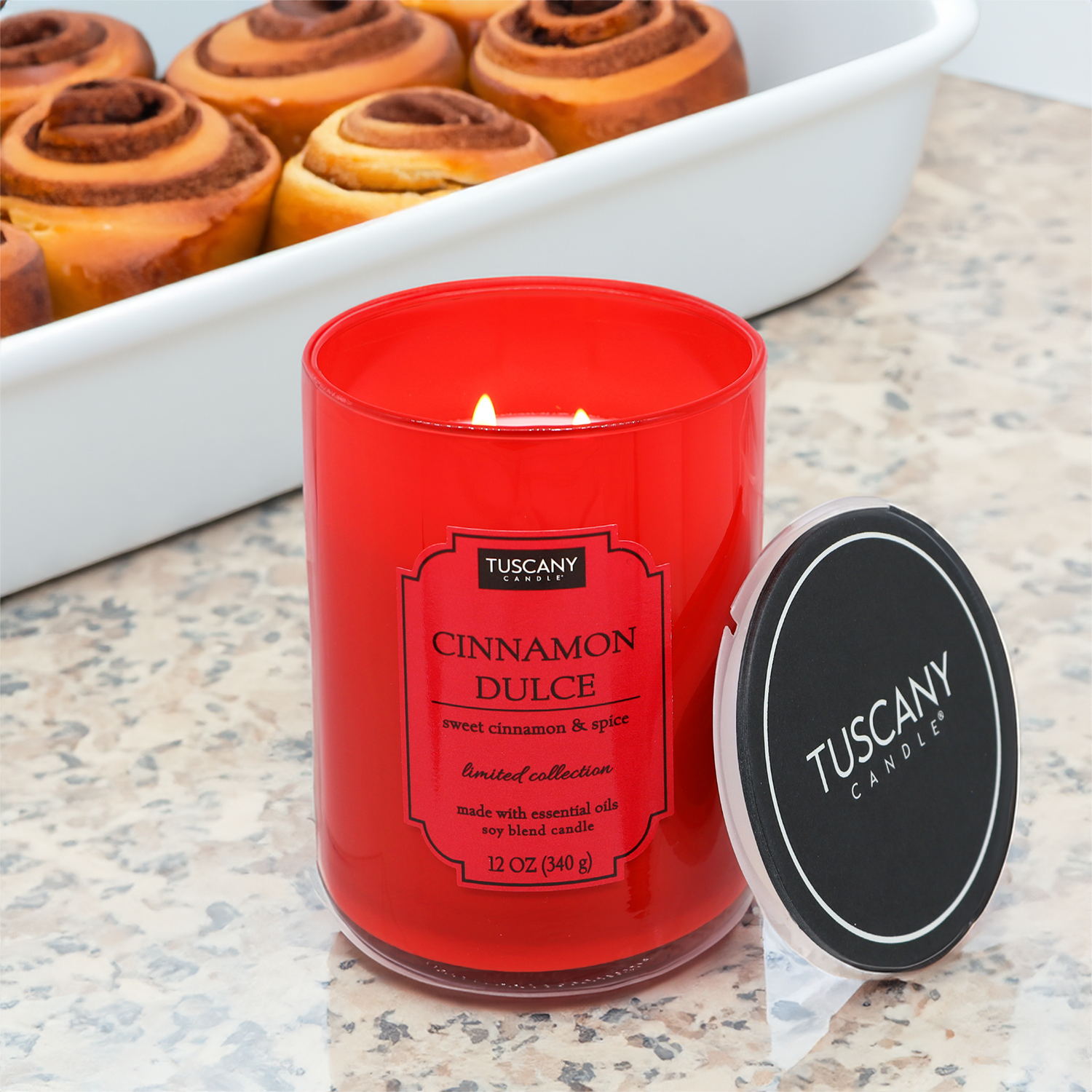 A Cinnamon Dulce candle from the Tuscany Candle® EVD Colorsplash collection next to a tray of sweet cinnamon and spice rolls.