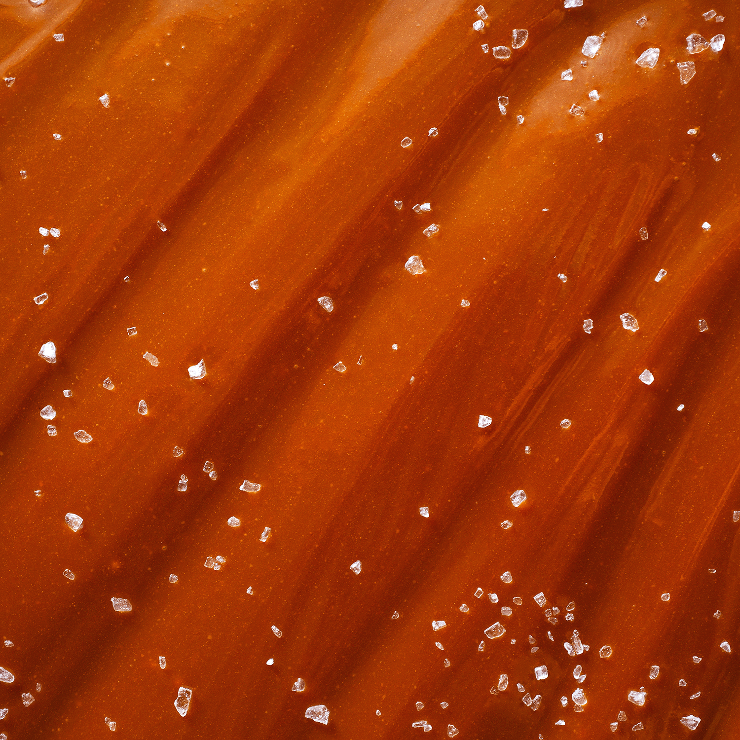 Close-up image of the Trick Or Treat Long-Lasting Scented Jar Candle (14 oz) by Tuscany Candle® SEASONAL, capturing the rich, smooth caramel hues of its wax and a sprinkling of coarse sea salt crystals on its surface, reminiscent of delectable toffee caramels.