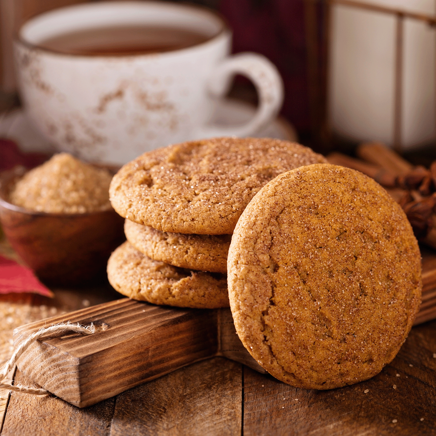 Three ginger cookies stacked on a wooden surface beside a whole cookie, a bowl of brown sugar, and a mug of tea in the background evoke the comforting snickerdoodle scent reminiscent of your favorite Tuscany Candle® SEASONAL Only Treats Scented Wax Melt (2.5 oz)—a perfect flameless alternative for cozy moments.