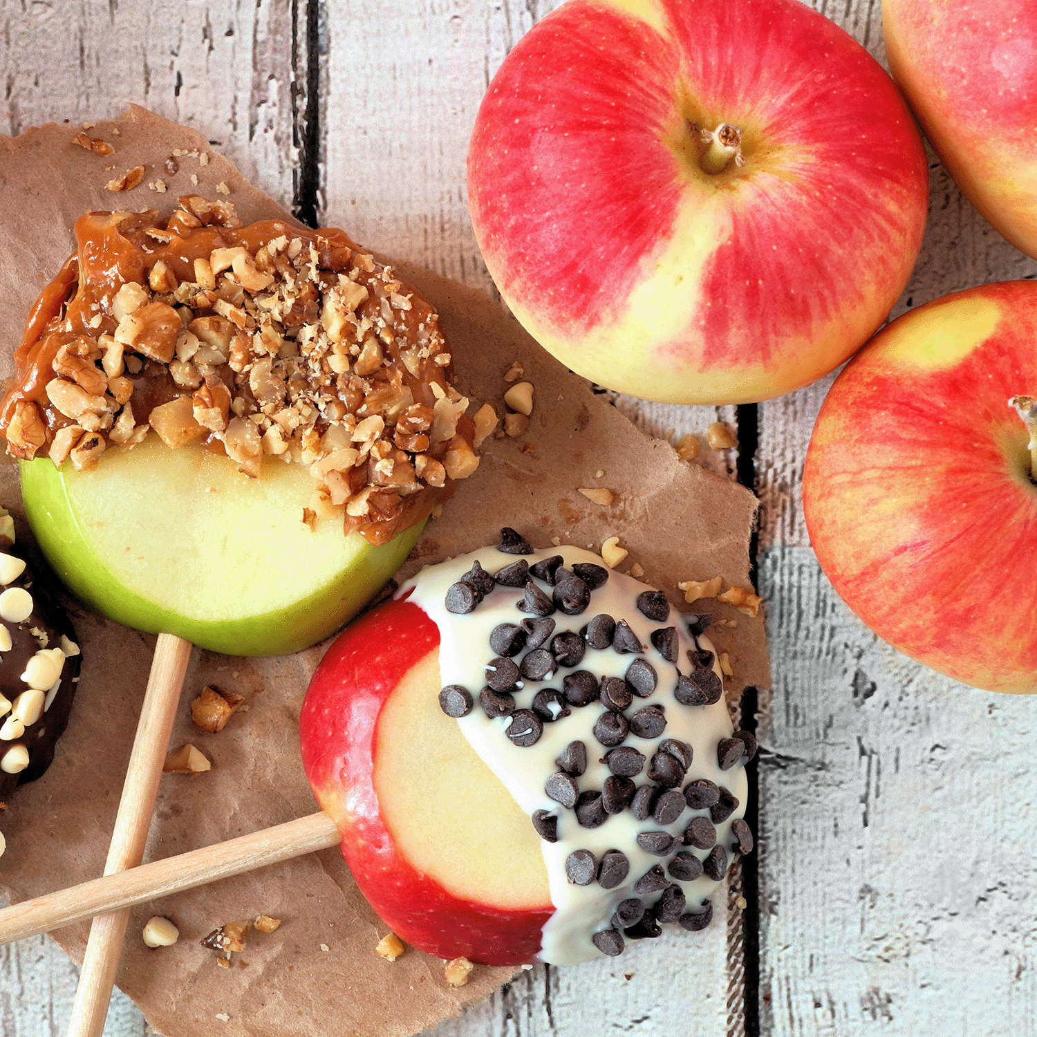 Three apples are coated in various toppings: one with caramel and nuts, one with chocolate and nuts, and one with white chocolate and chocolate chips. Whole apples are also present in the background, evoking a scene reminiscent of the Tuscany Candle® SEASONAL Bewitching Brew Scented Wax Melt collection.