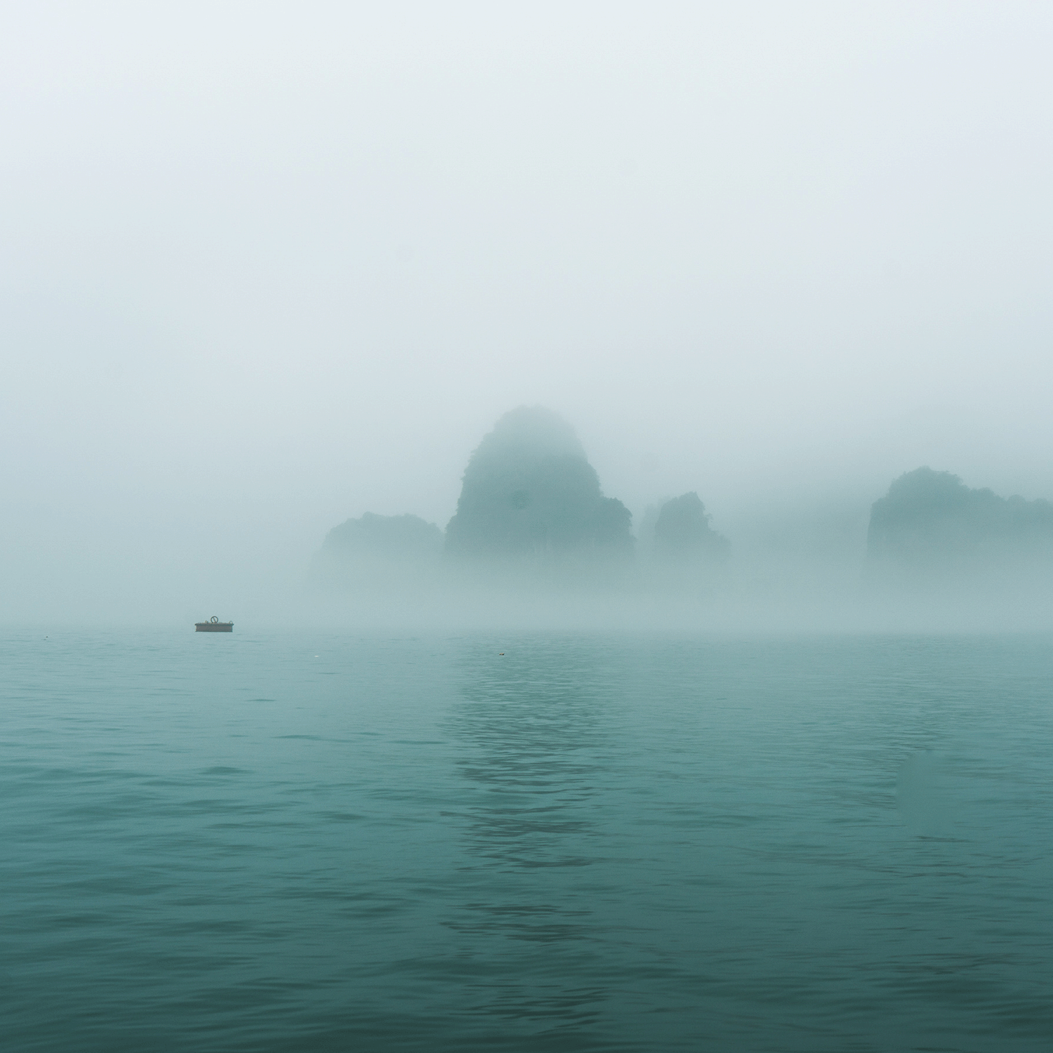 A small boat floats on calm water with dreamy mist-shrouded rocky islands in the background, encapsulating the serene essence of the Tear Drops Of A Mermaid Long-Lasting Scented Jar Candle (18 oz) from Tuscany Candle® SEASONAL's Apothecary Collection.