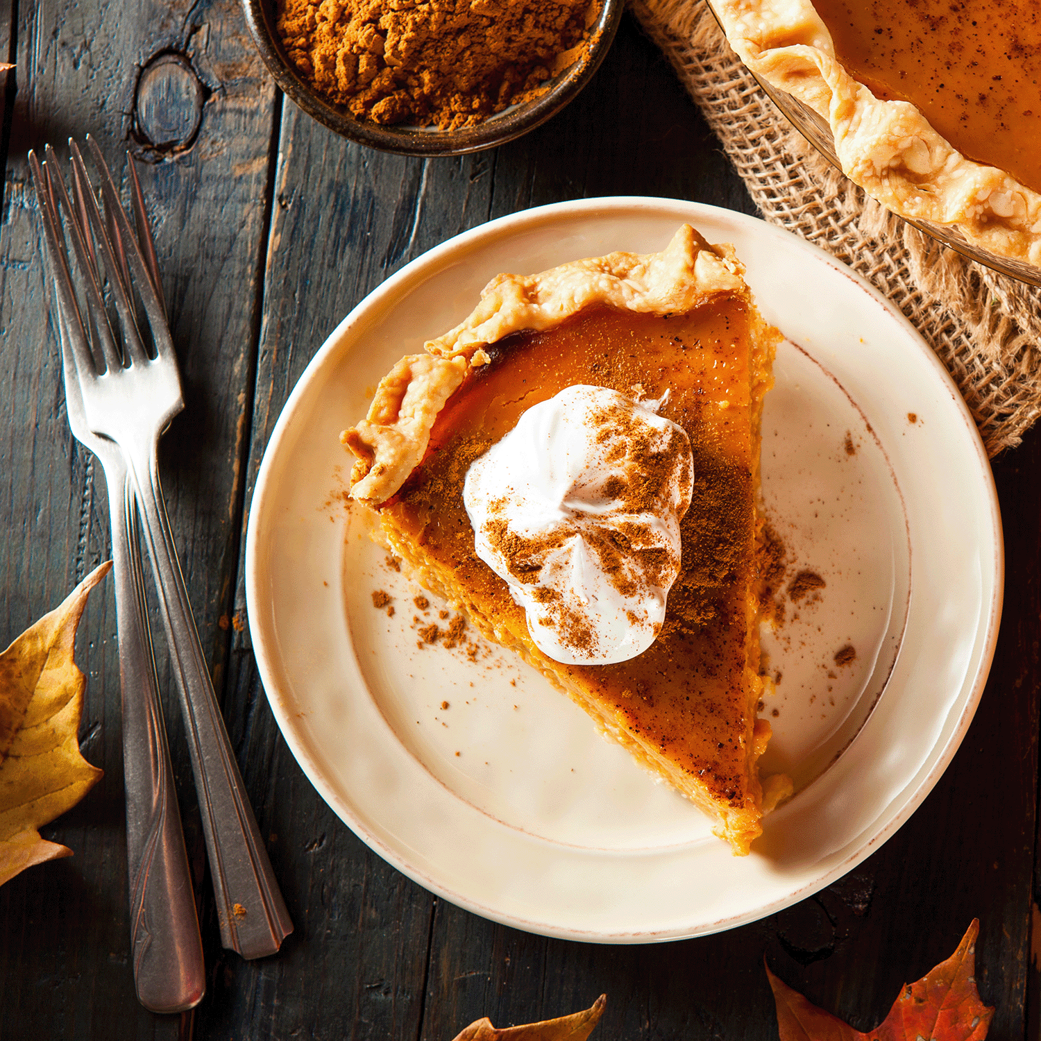 A slice of pumpkin pie topped with whipped cream sits on a plate with cinnamon dusting. Forks are on the side, and fall leaves decorate the rustic wooden table, filling the air with cozy autumnal aromas reminiscent of Tuscany Candle® SEASONAL's Spooky Pumpkin Spice Long-Lasting Scented Jar Candle (12 oz).