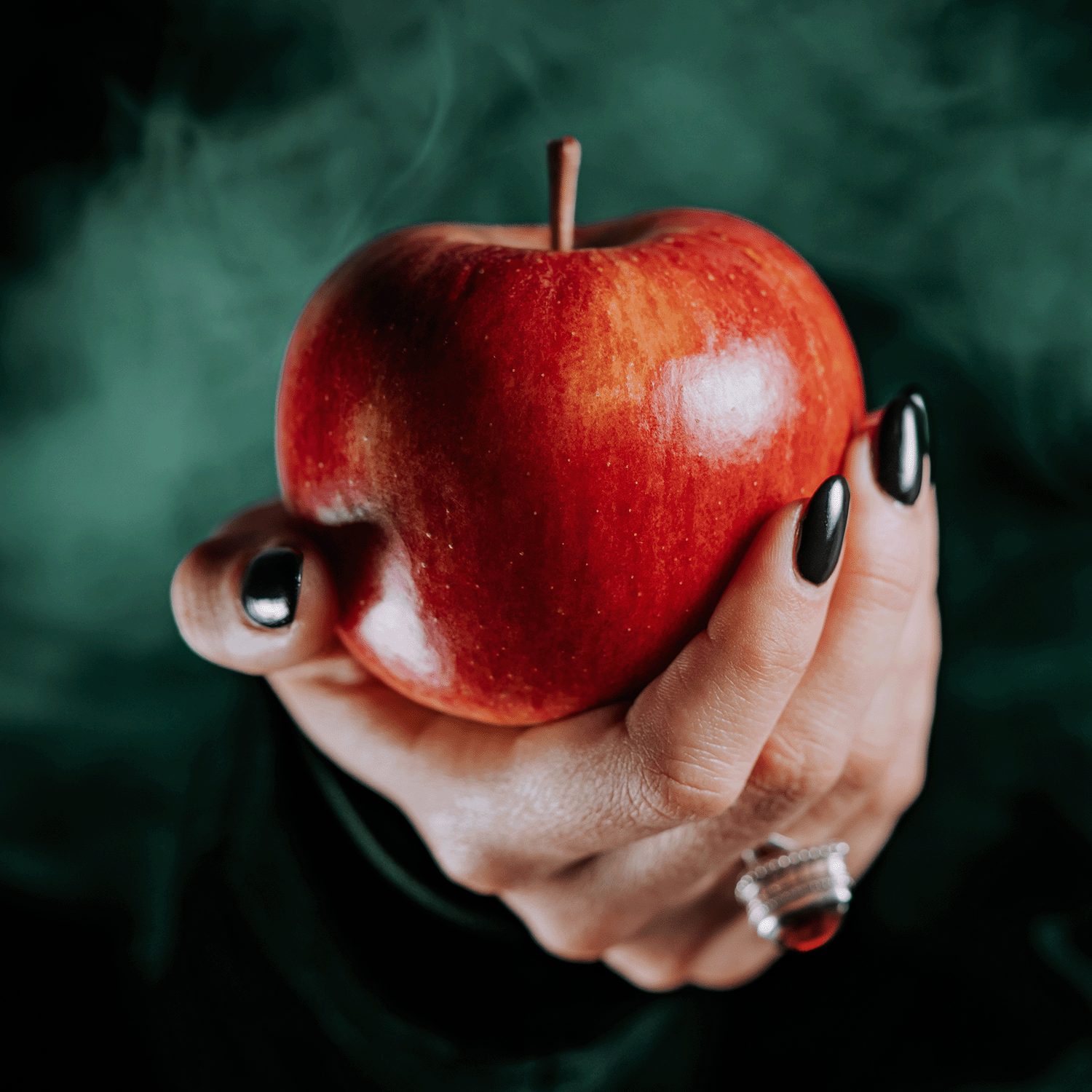 A hand adorned with black nail polish holds a shiny red apple against a smoky dark background, evoking the fragrant aroma of the Bobbing For Apples Long-Lasting Scented Jar Candle (12 oz) by Tuscany Candle® SEASONAL.